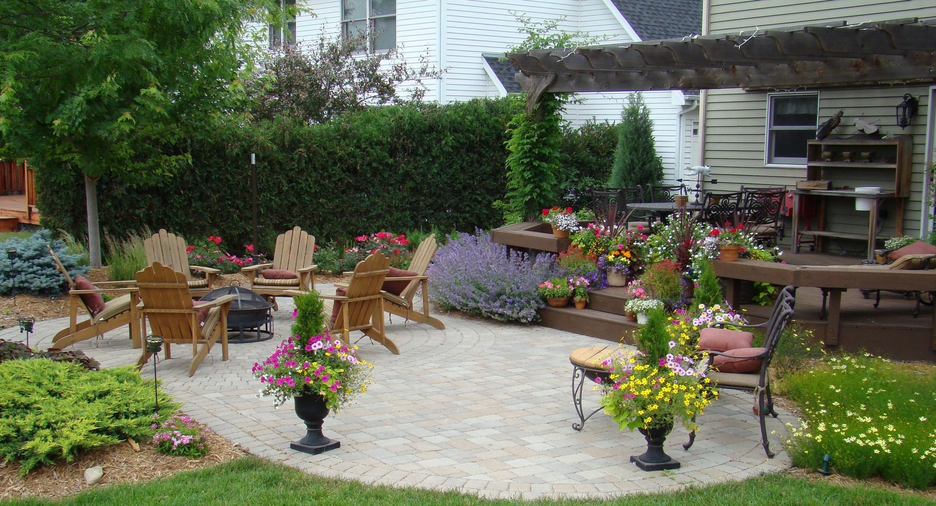 A backyard patio with Adirondack chairs around a fire pit, surrounded by plants and a pergola.