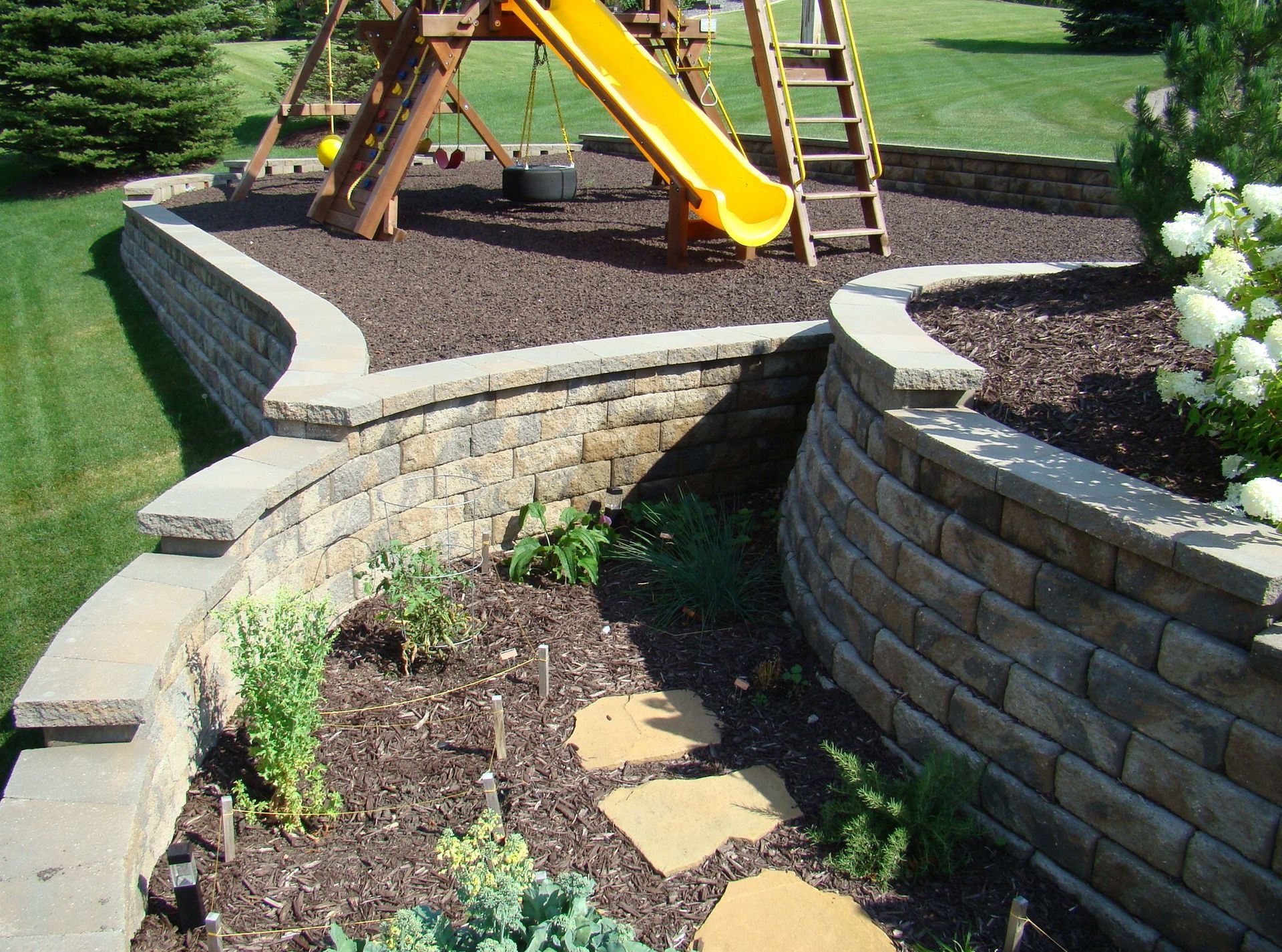 Stone retaining walls frame a playground, garden, and walkway on a grassy lawn.