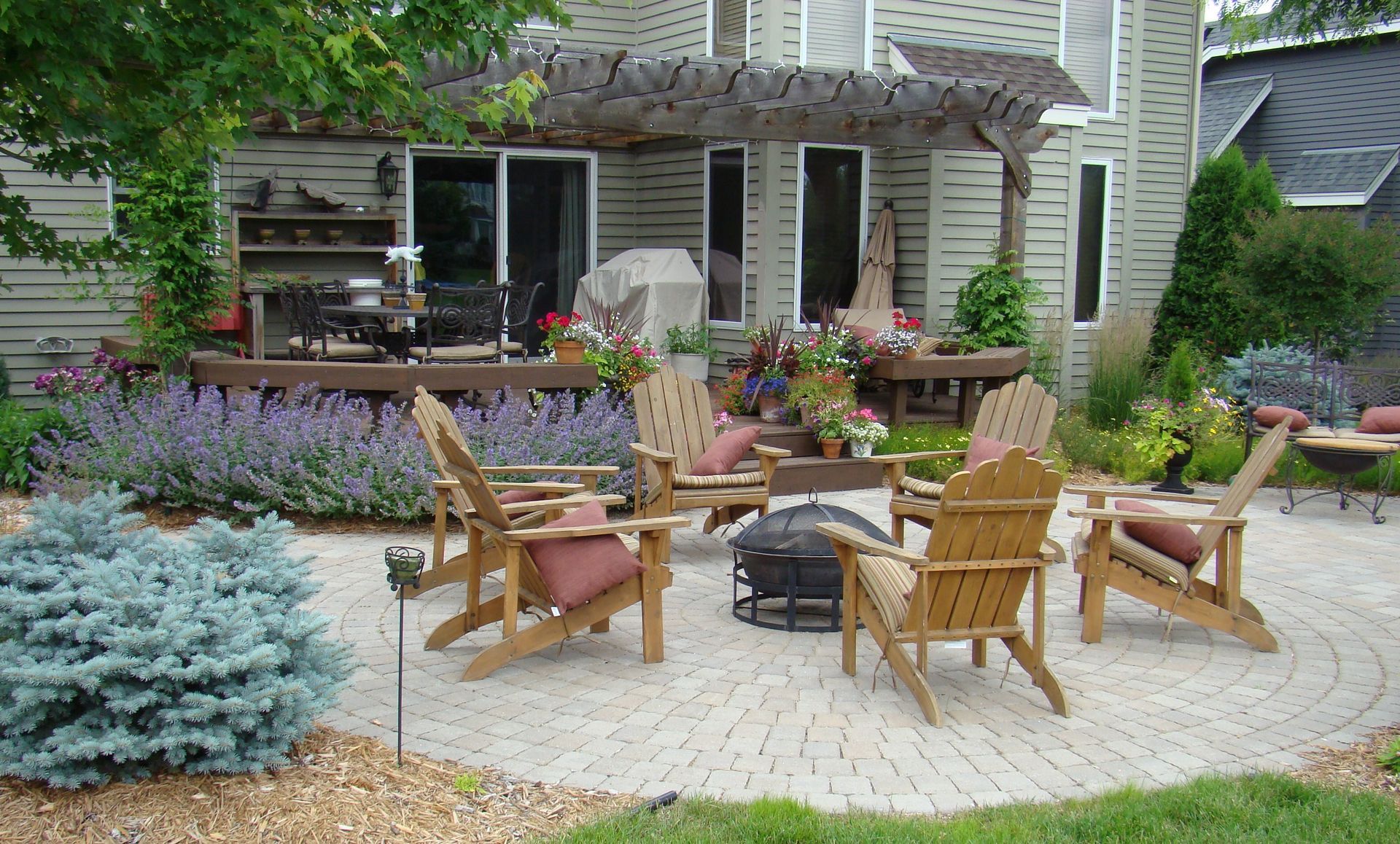 Backyard patio with wooden chairs, fire pit, and flowering plants.