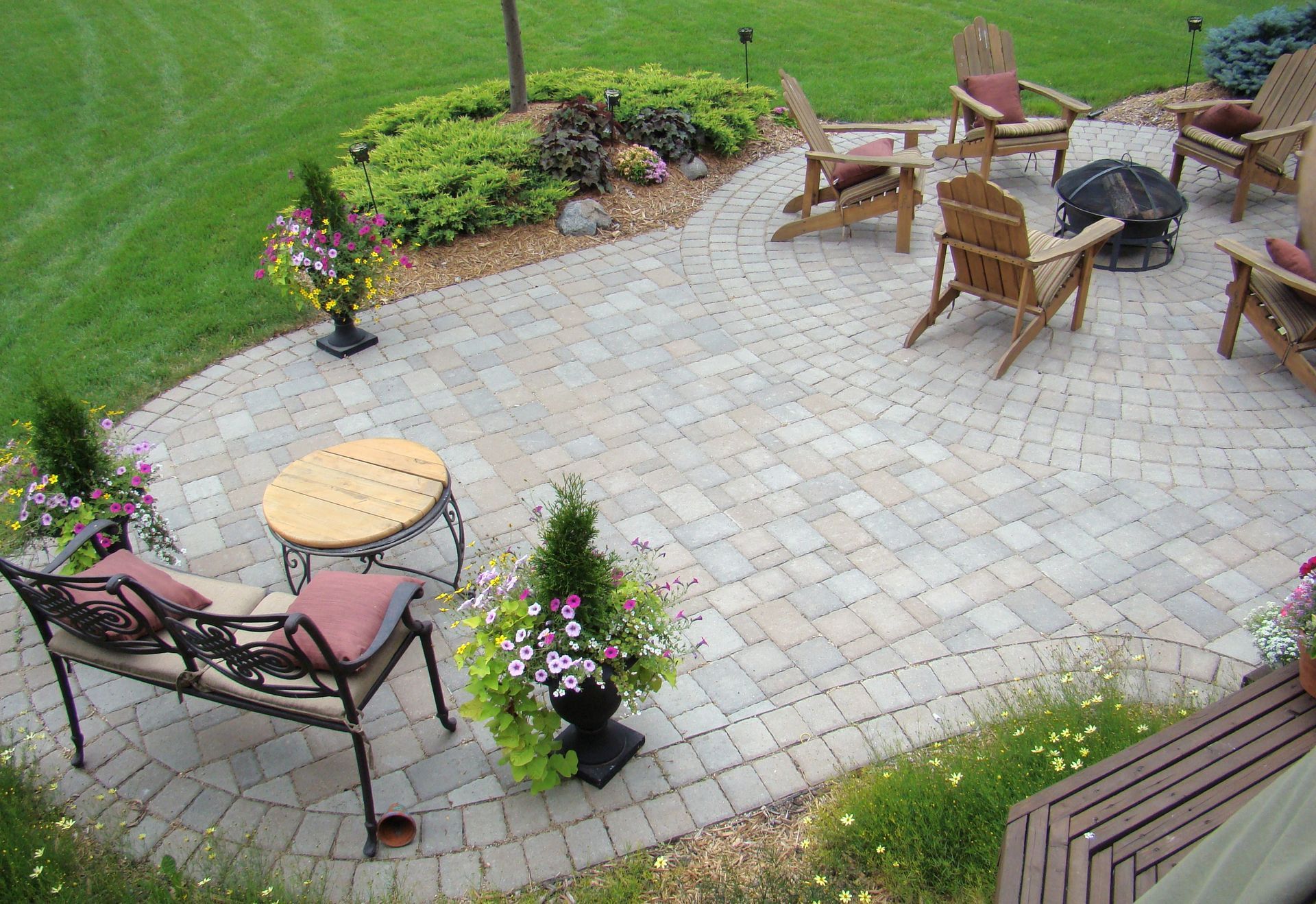 Stone patio with seating area, fire pit, and potted plants, surrounded by green grass.