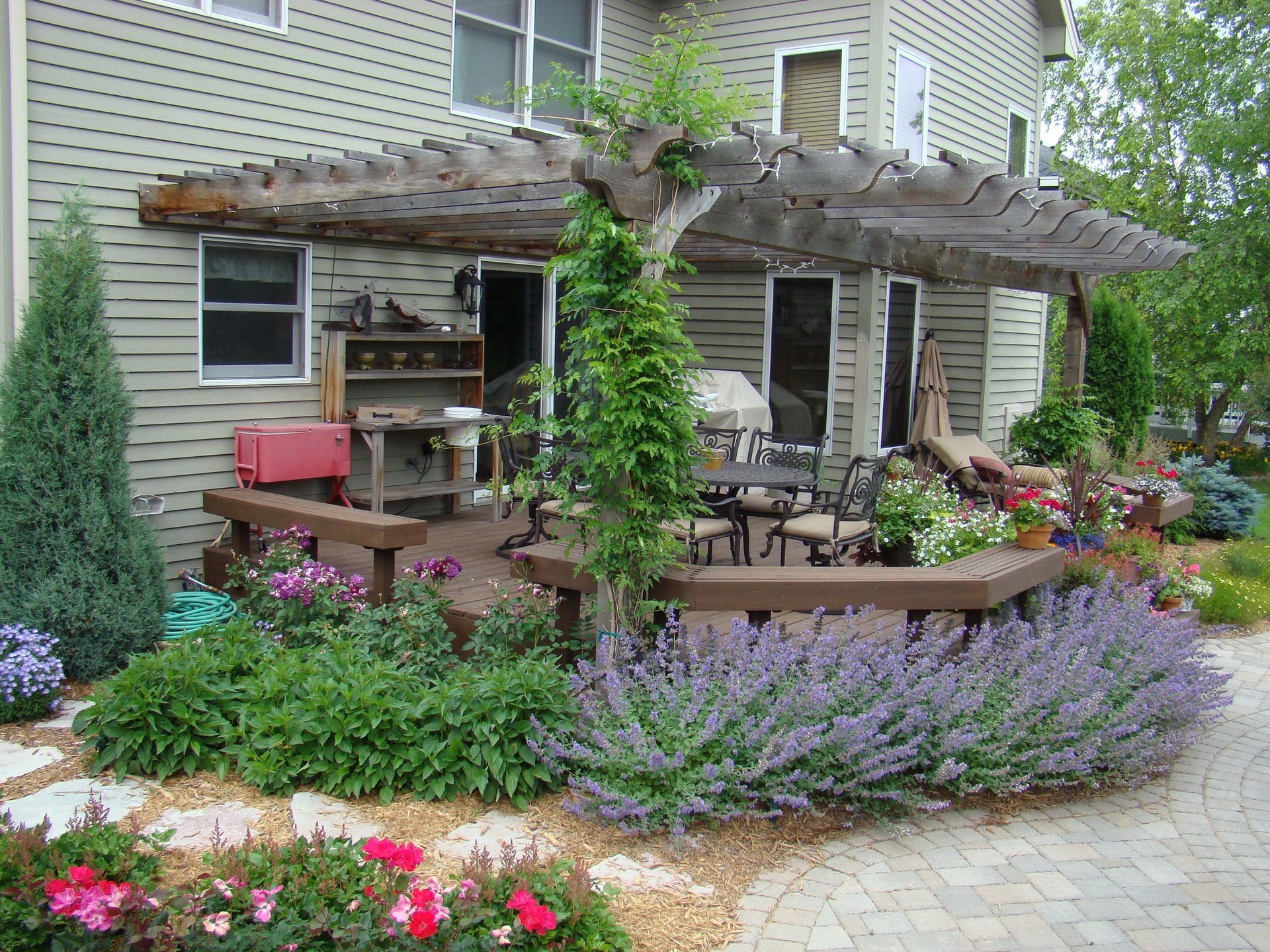 Backyard deck with pergola, surrounded by flowerbeds. Patio furniture under pergola, house in the background.