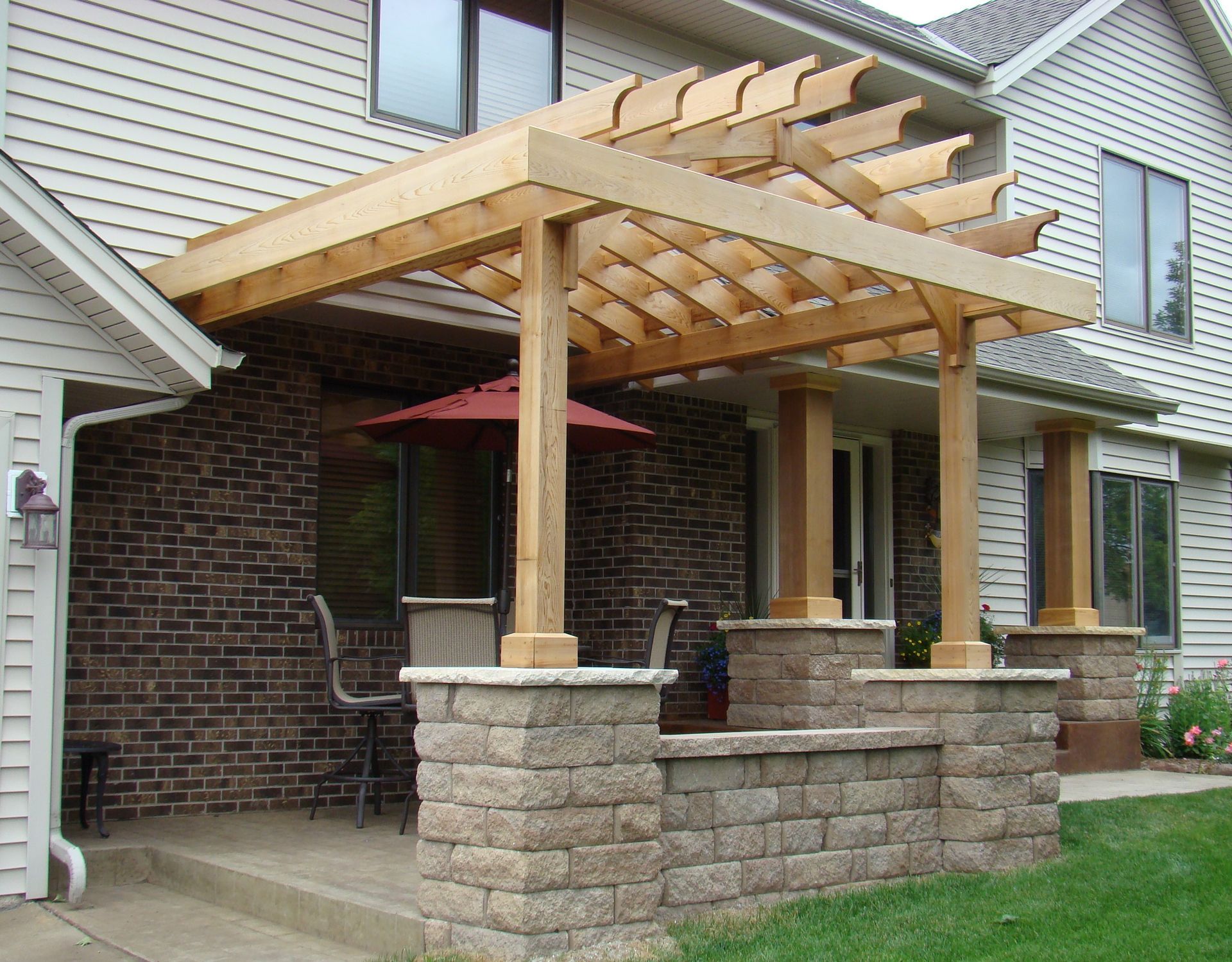 Wooden pergola over a brick patio with stone pillars, a red umbrella, and seating.