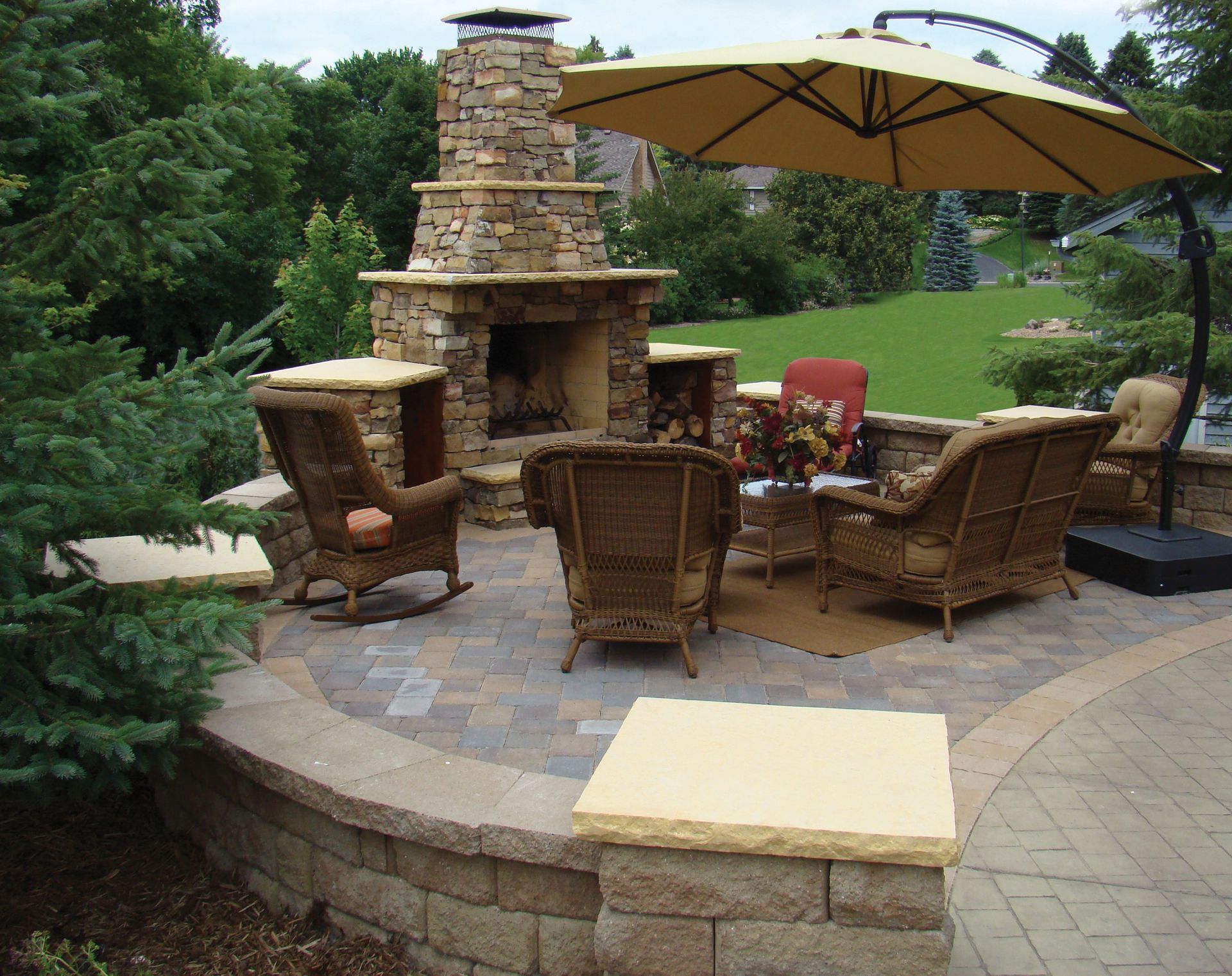 Outdoor patio with stone fireplace, wicker chairs, umbrella, and surrounding greenery.
