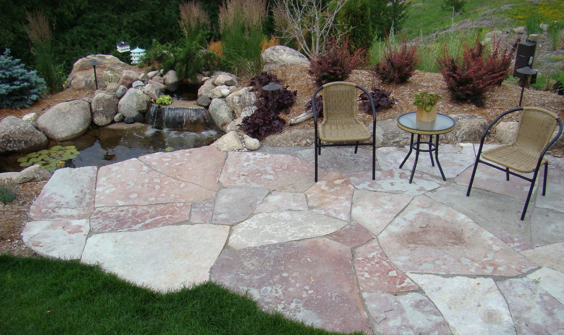 Stone patio with pond, waterfall, and seating area. Red and beige flagstones, brown wicker chairs, and greenery.