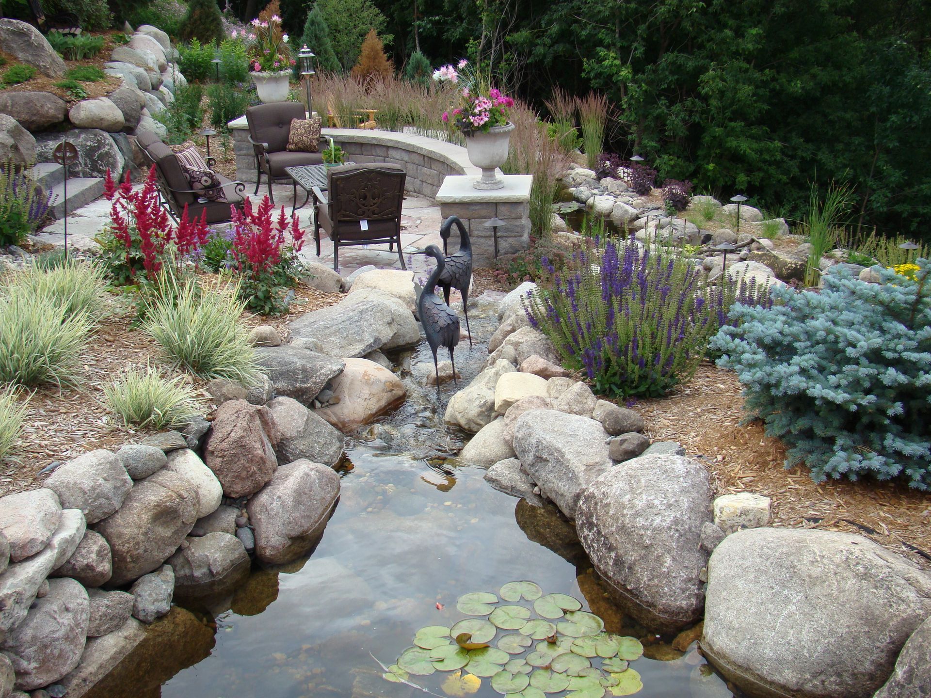 Stone-lined water feature with lush plants and seating area, two heron statues stand in the water.