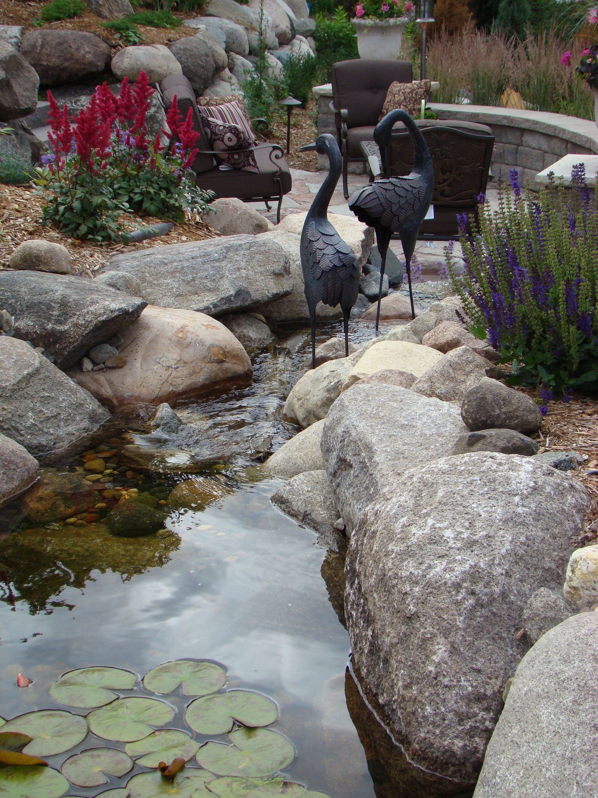 Water garden with granite boulders, metal crane sculptures, and flowering plants.