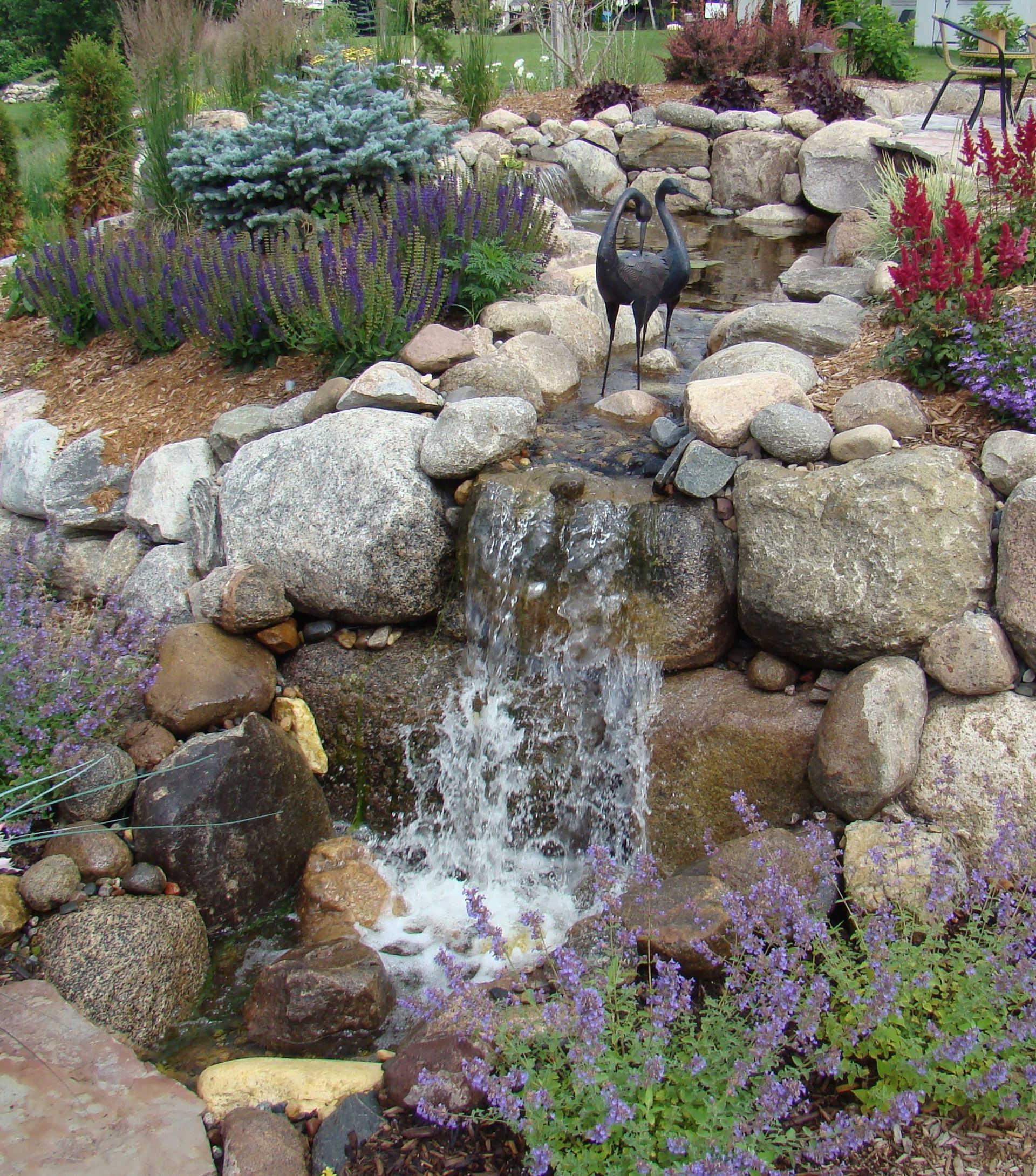 Waterfall feature in a landscaped garden with layered rocks, flowers, and a black statue.