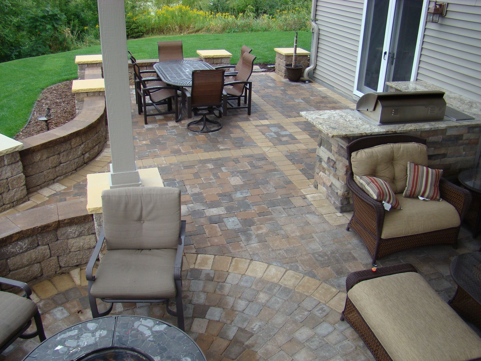 Patio with stone pavers, seating areas, outdoor kitchen, dining table, and green lawn.