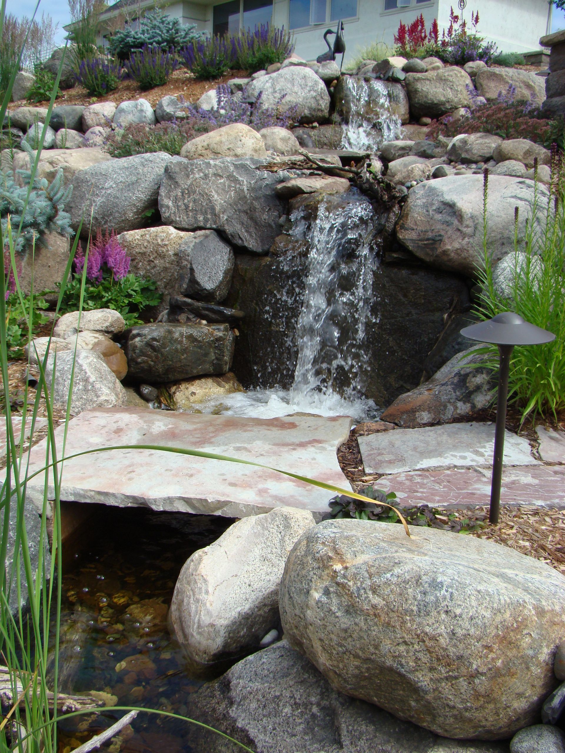 Waterfall cascading over large rocks into a pond, with lush greenery and a stone bridge.