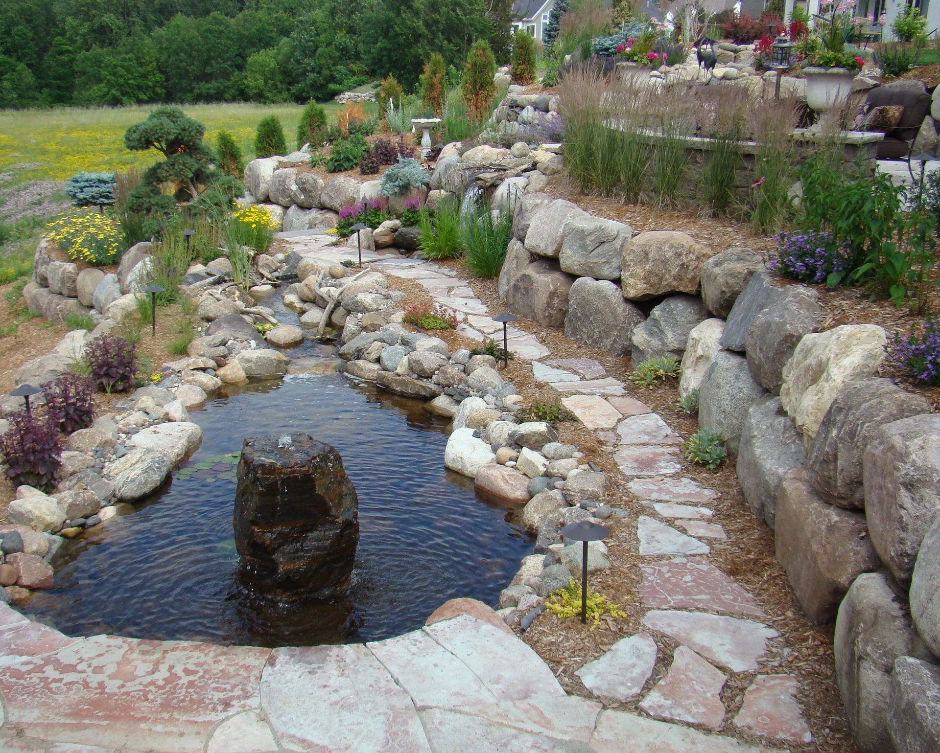 Landscaped water feature with pond, stone pathway, cascading stream, and various plants.