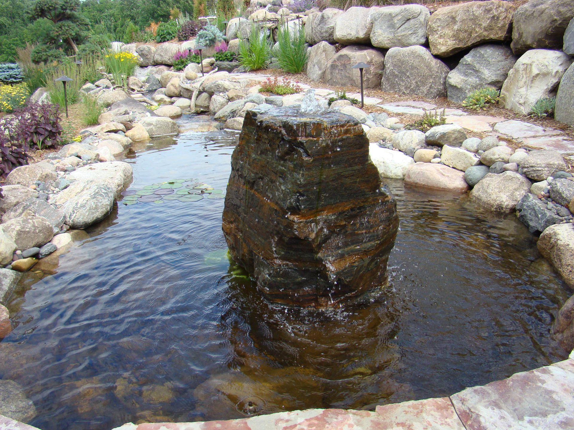 Water fountain in a stone-lined pond, surrounded by rocks and plants.