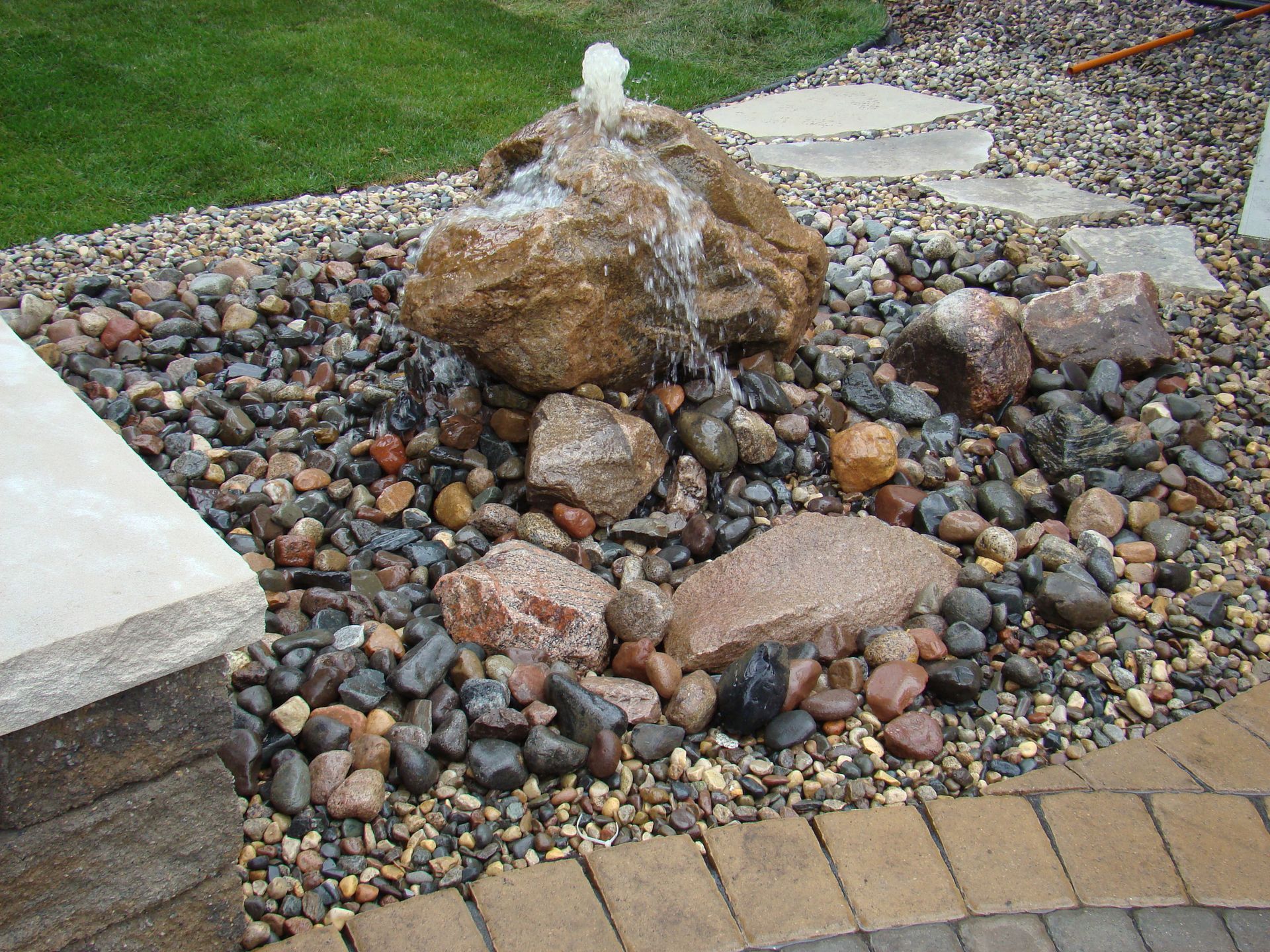 Rock water fountain in a bed of pebbles, with stone pavers and brick edging.