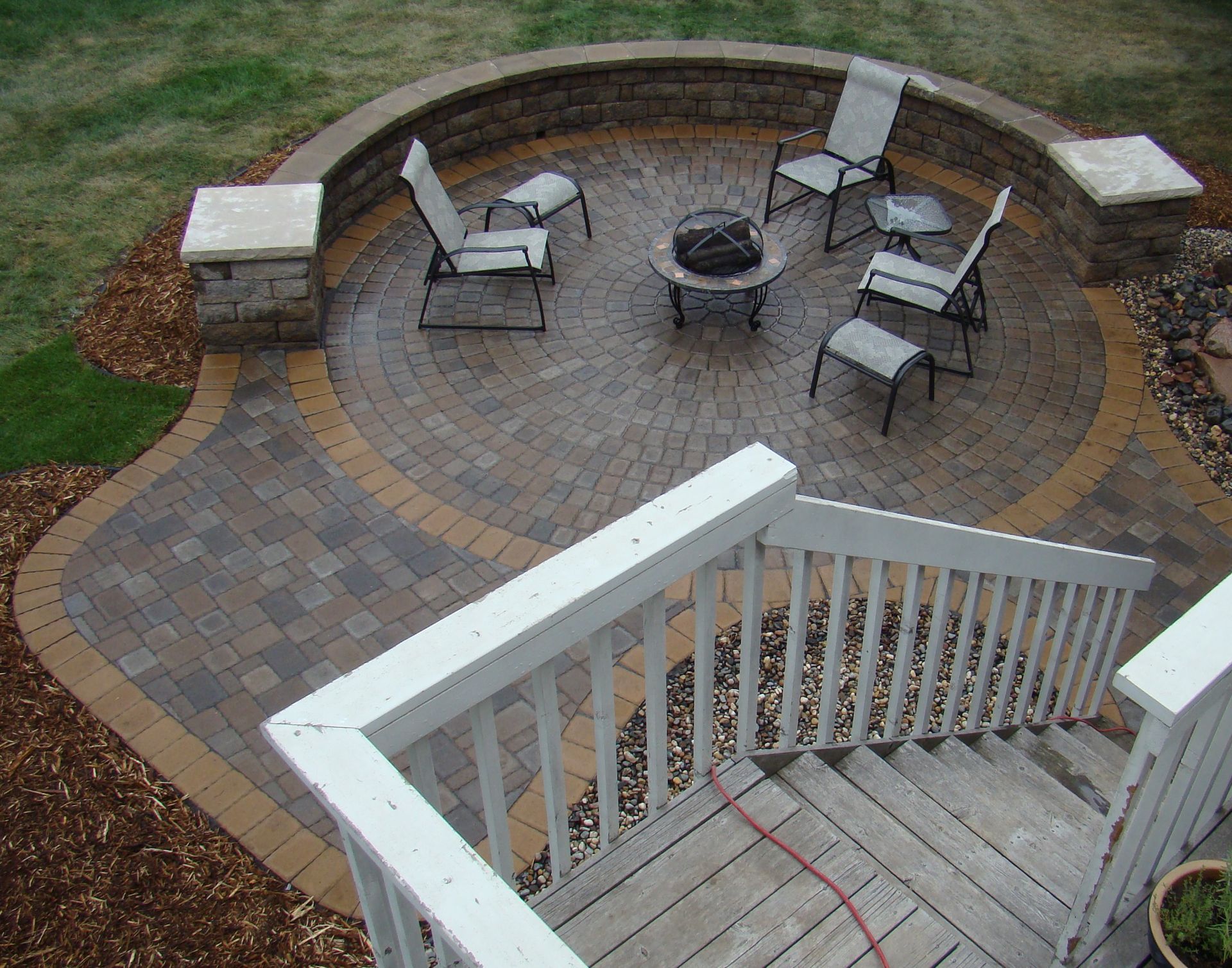 Circular brick patio with seating around a fire pit, bordered by a retaining wall and adjacent to a wooden deck.