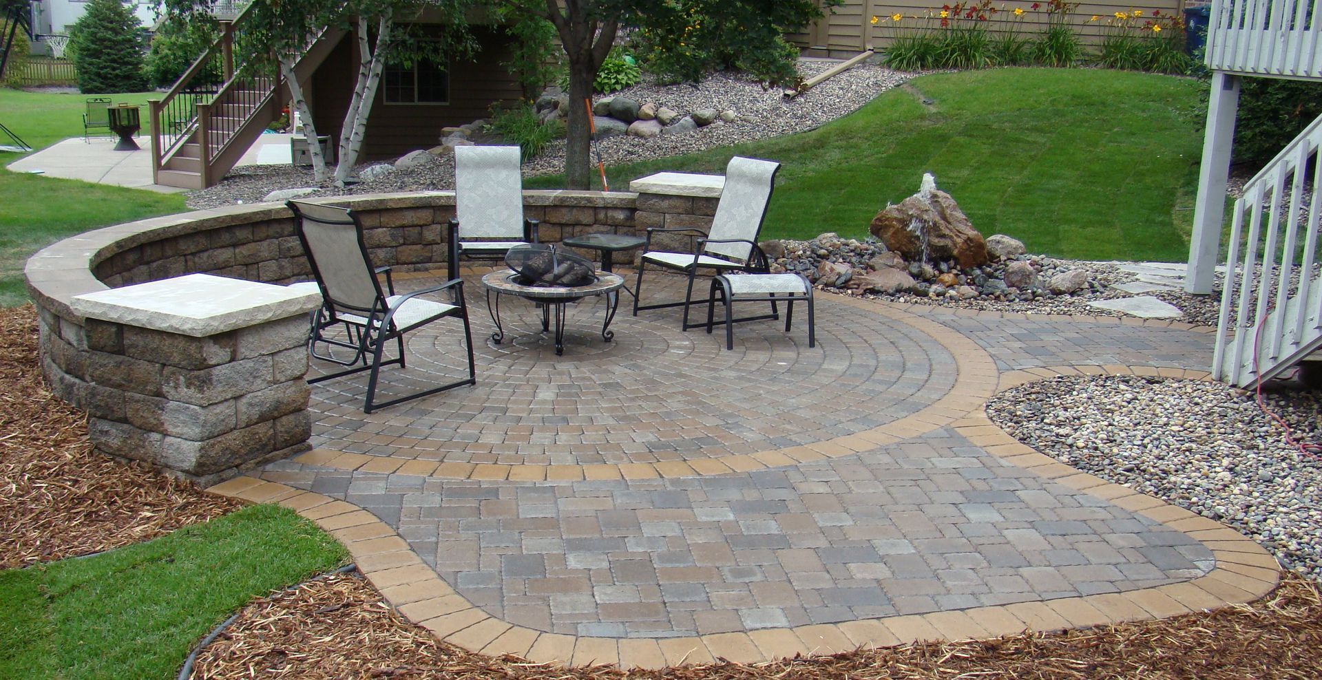 Brick patio with fire pit, chairs, and stone wall, in a landscaped backyard setting.