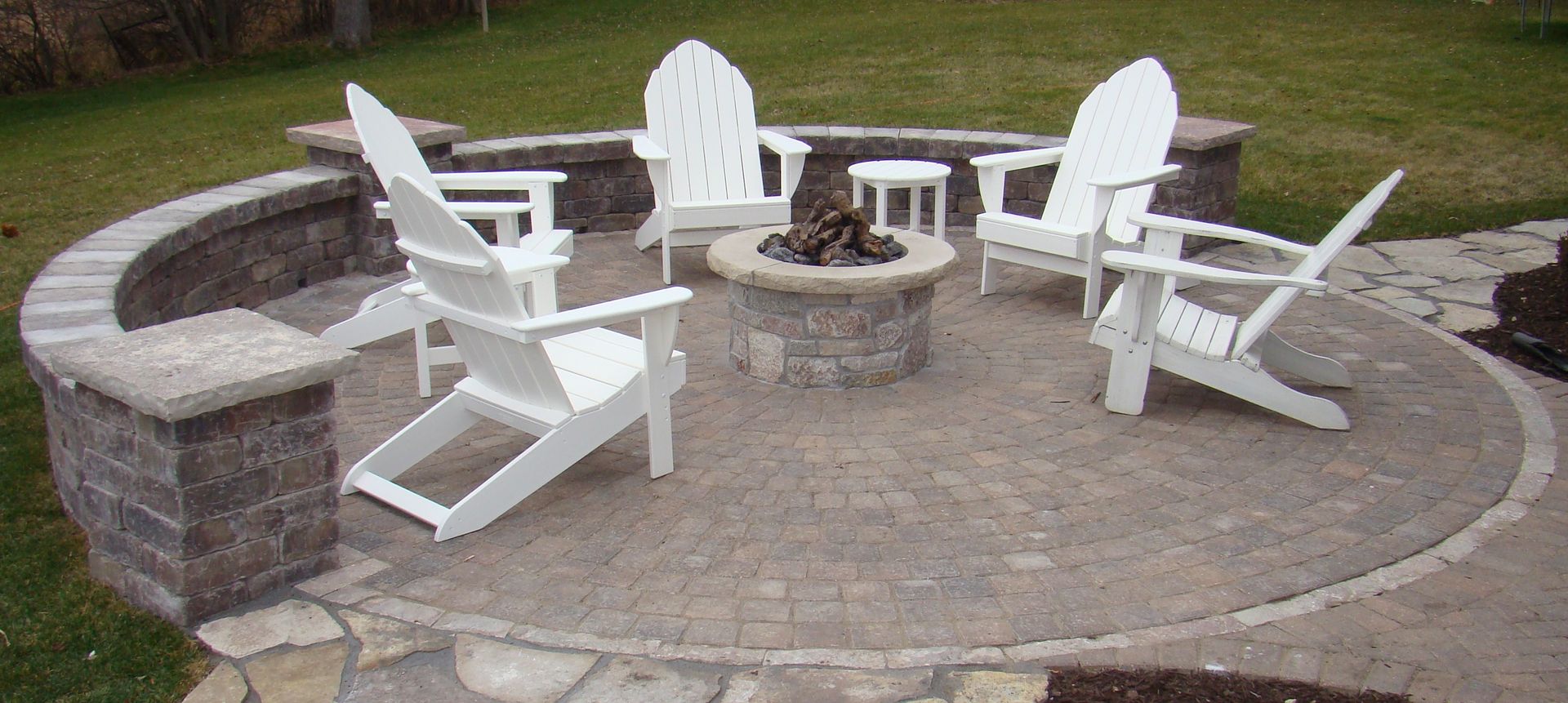 Outdoor fire pit with white Adirondack chairs arranged around it, on a stone patio.