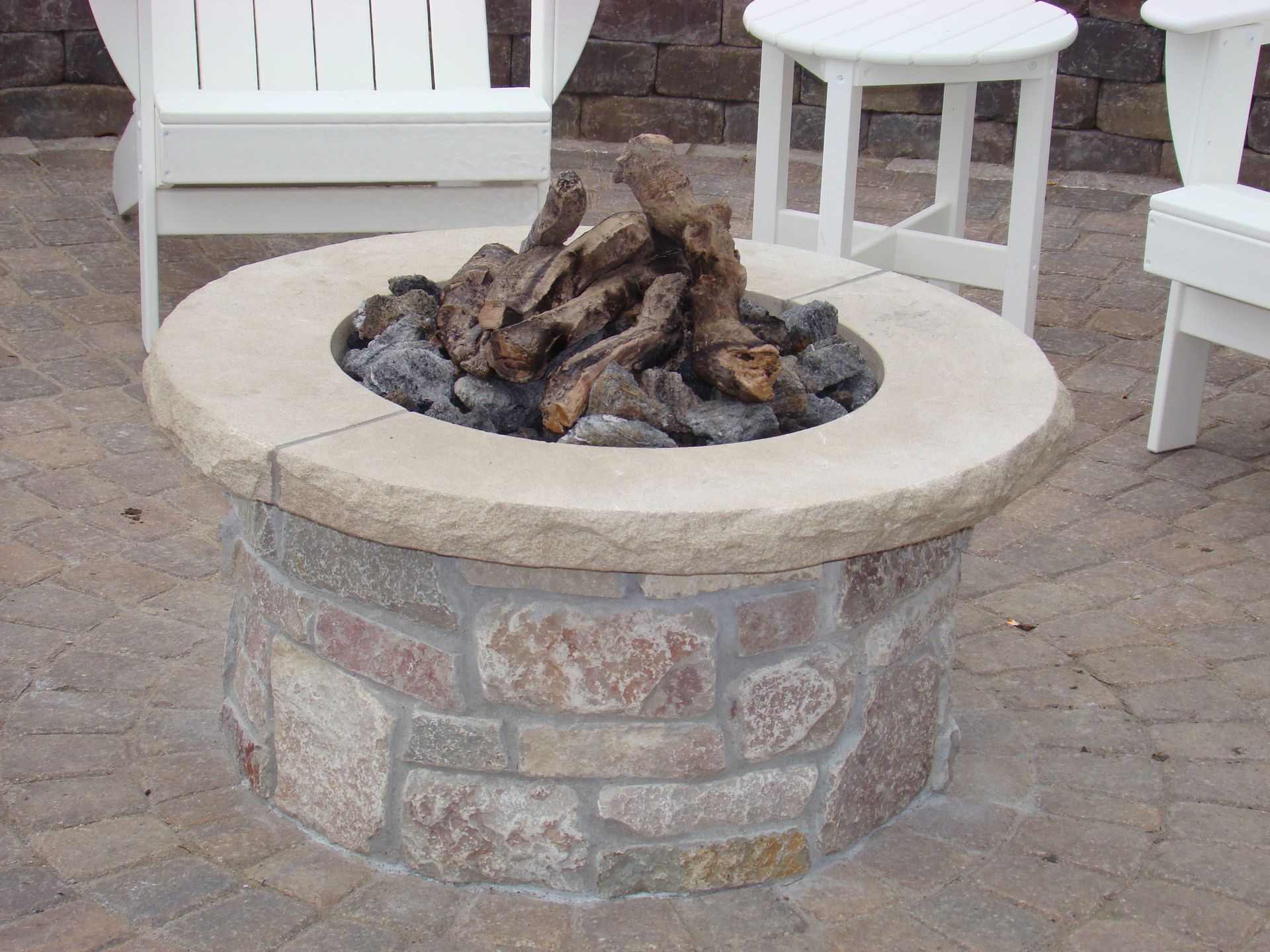 Stone fire pit with faux wood logs and surrounding chairs on a brick patio.