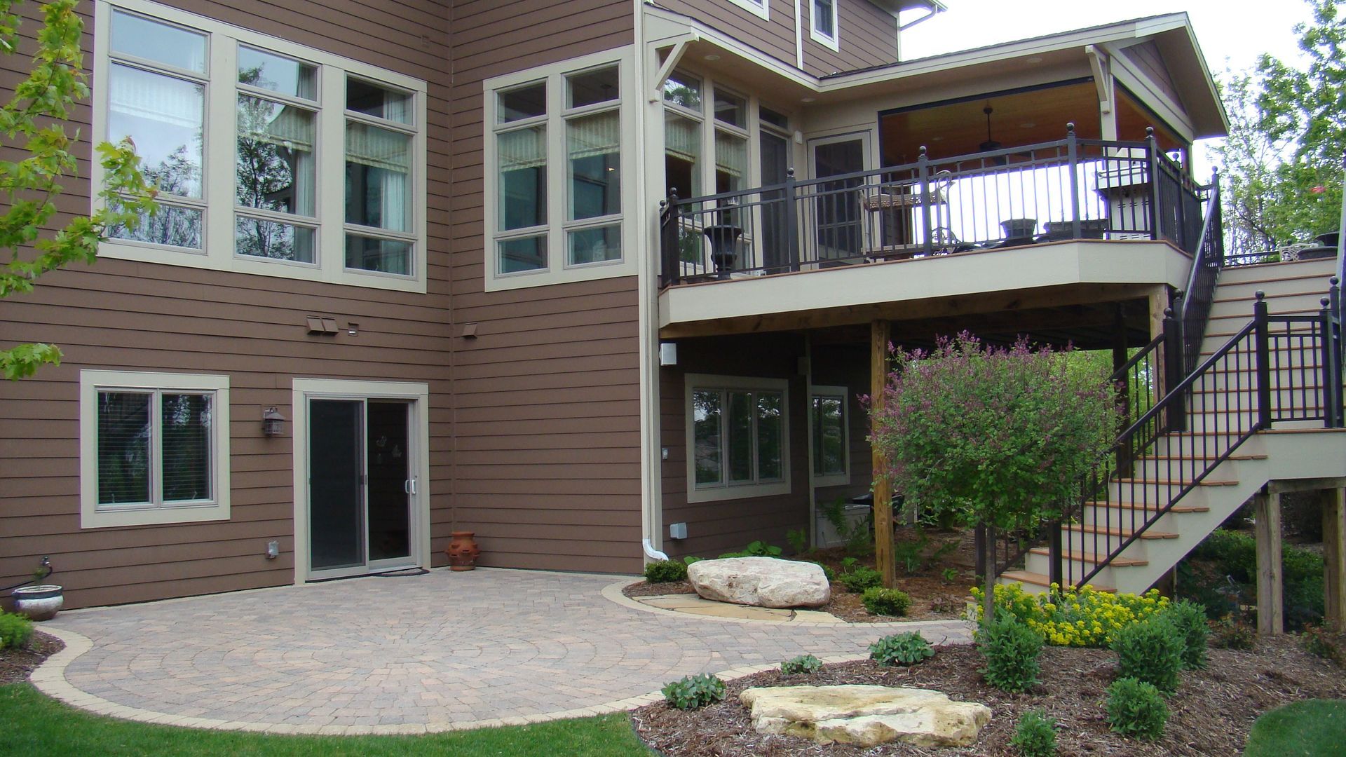 Brown house with a patio and elevated deck, stairs, and landscaping.