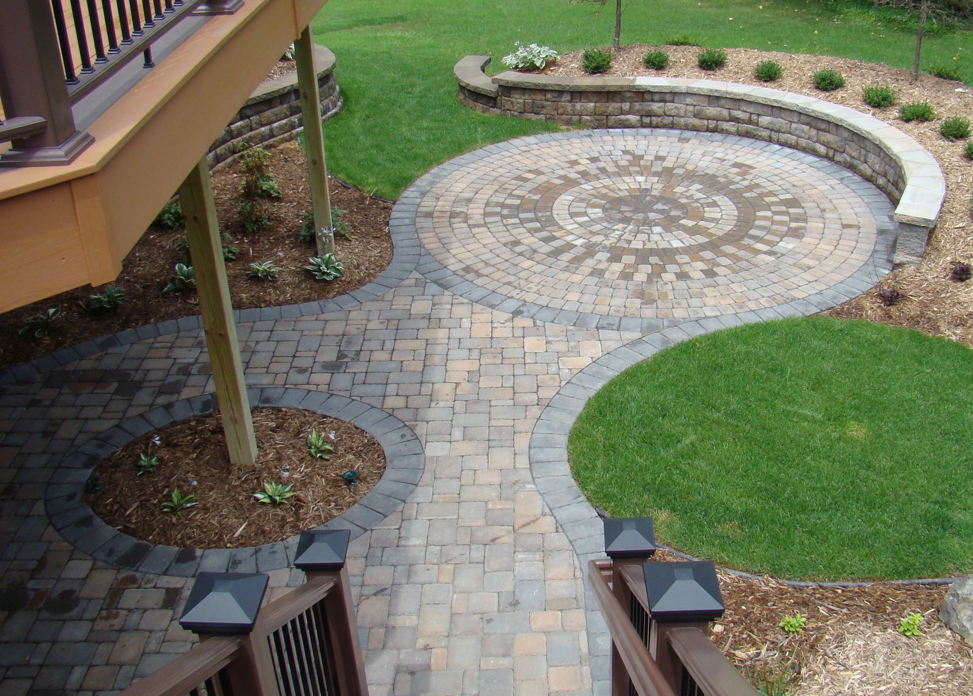 Stone patio with walkway leading to deck stairs and circular lawn features.