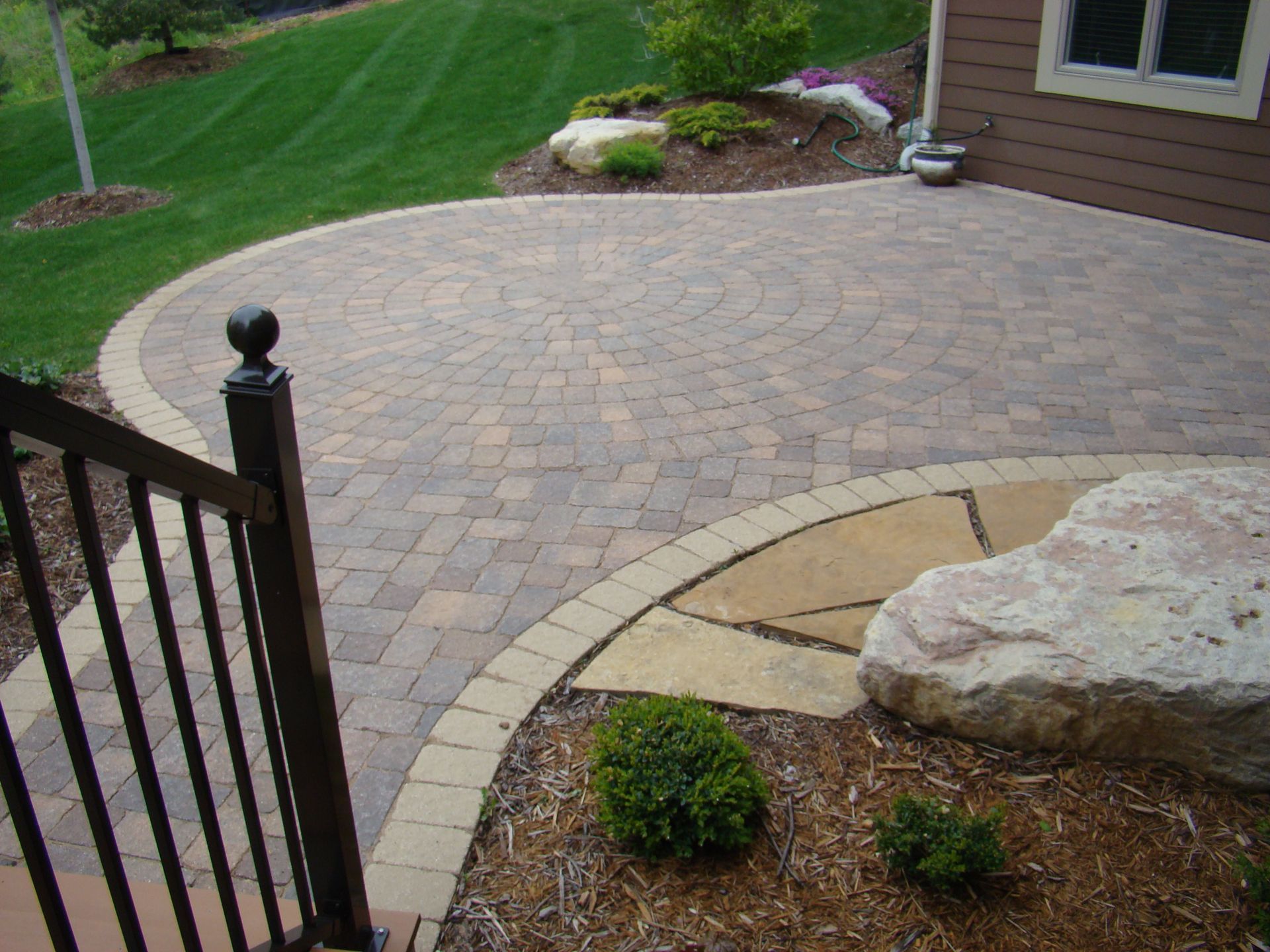 Paver patio with curved design, bordering a lawn, rock, and building.