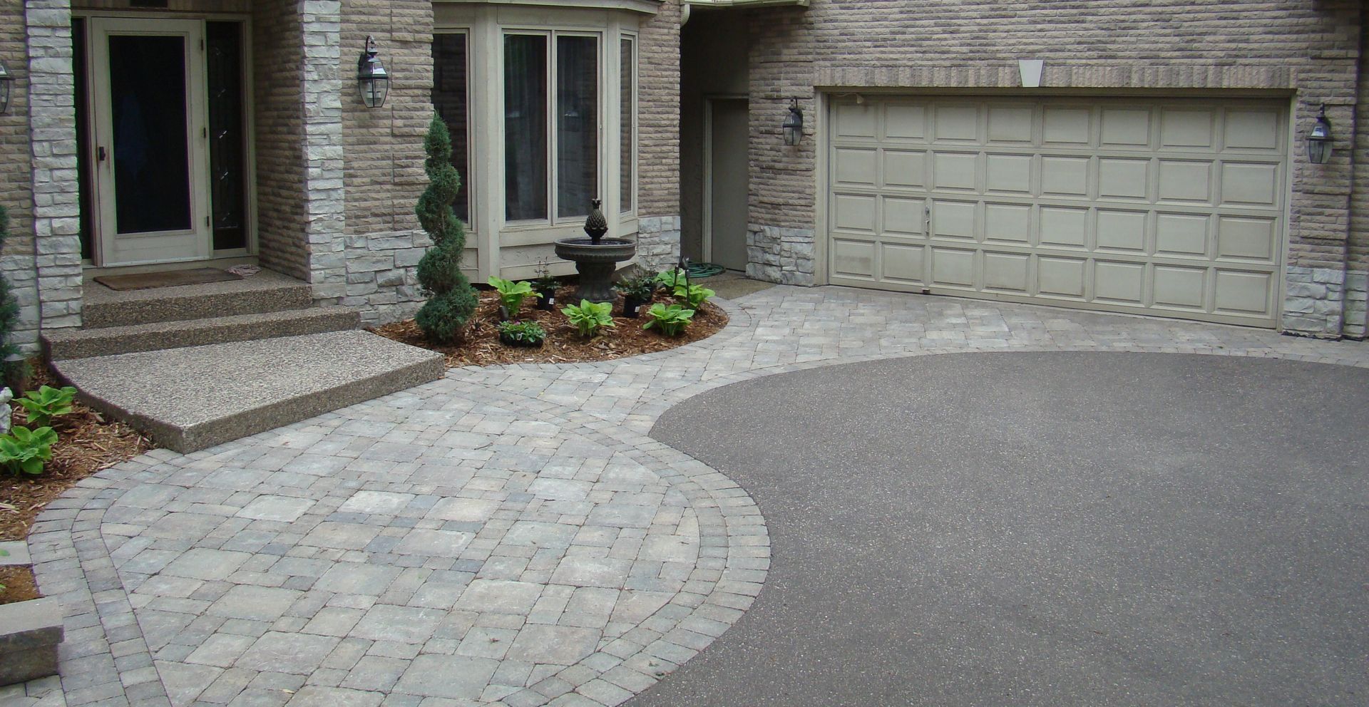 Brick home exterior with patterned paver walkway, garden, and driveway leading to a garage.