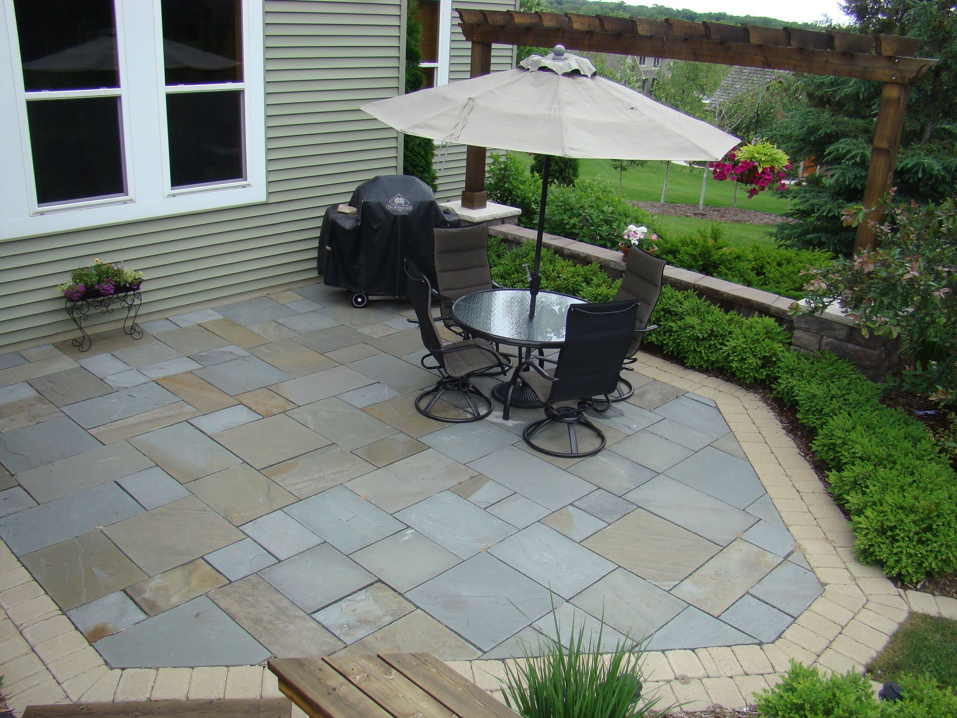 Patio with stone pavers, table, chairs, umbrella, grill, and lush greenery.