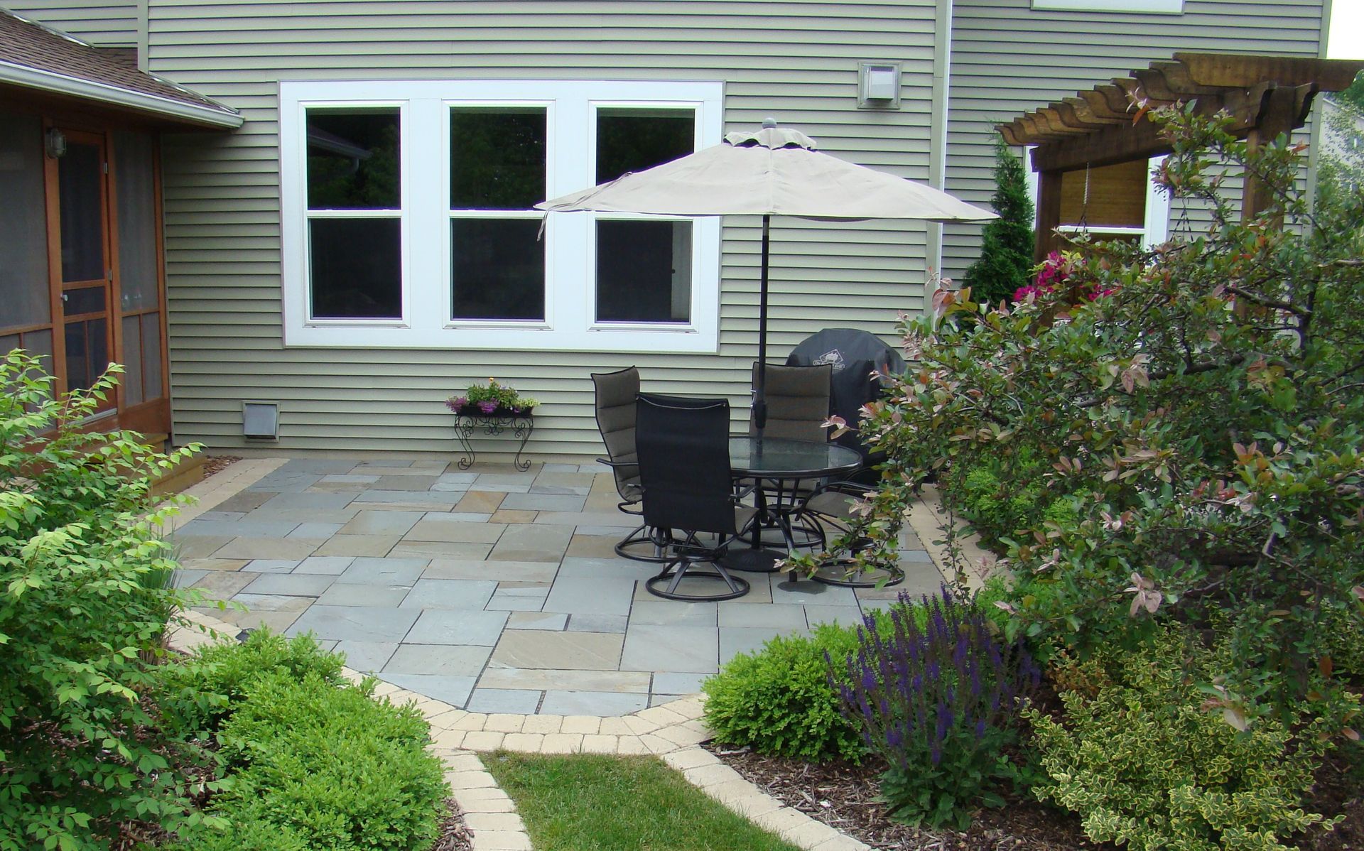 Patio with dining set, umbrella, and grill surrounded by greenery and the house.