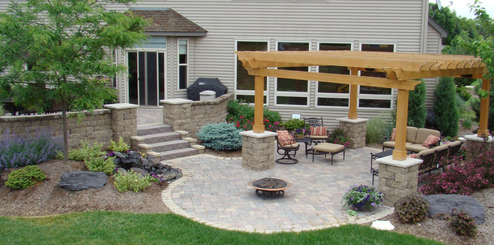 Patio with pergola, seating, fire pit, stone walls, landscaping, and a house in the background.