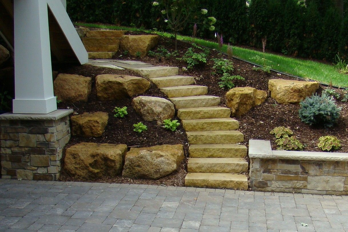 Stone steps and retaining wall leading up a slope with large boulders and plantings.
