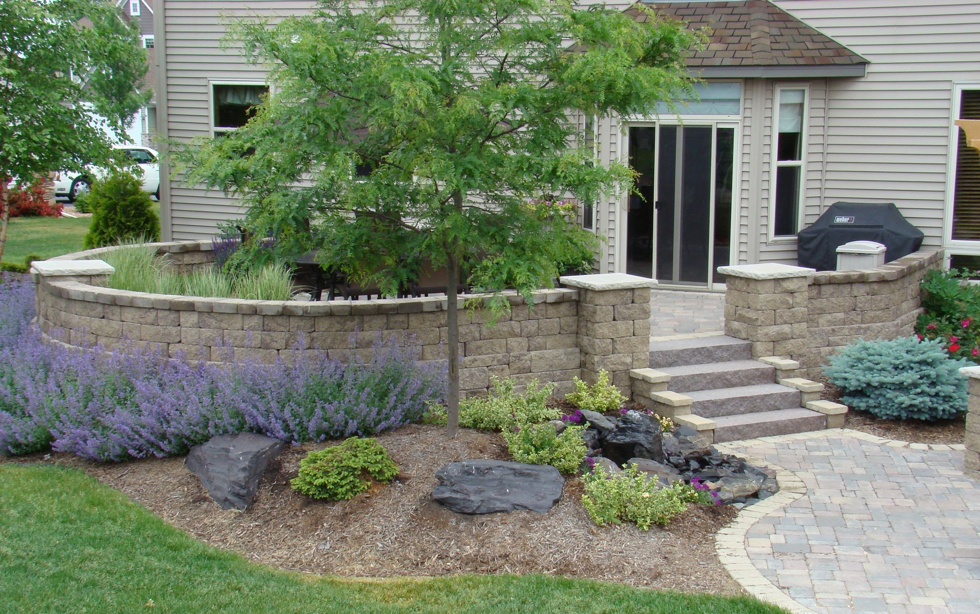 Landscaped backyard with stone walls, steps, patio, tree, and purple flowers.