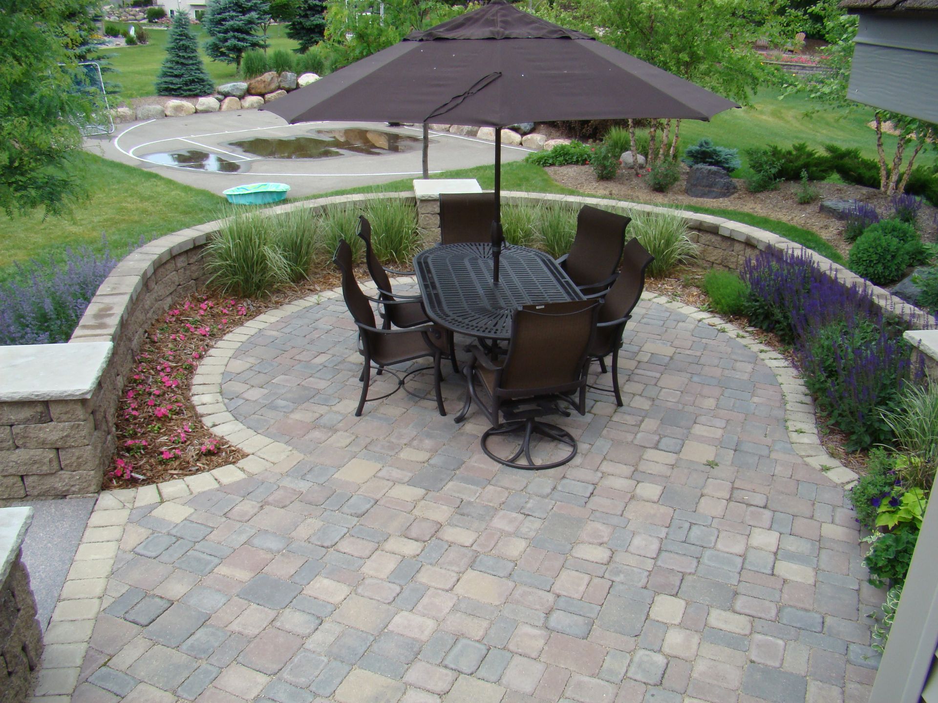 Patio with outdoor dining set under umbrella, surrounded by landscaping and retaining walls.