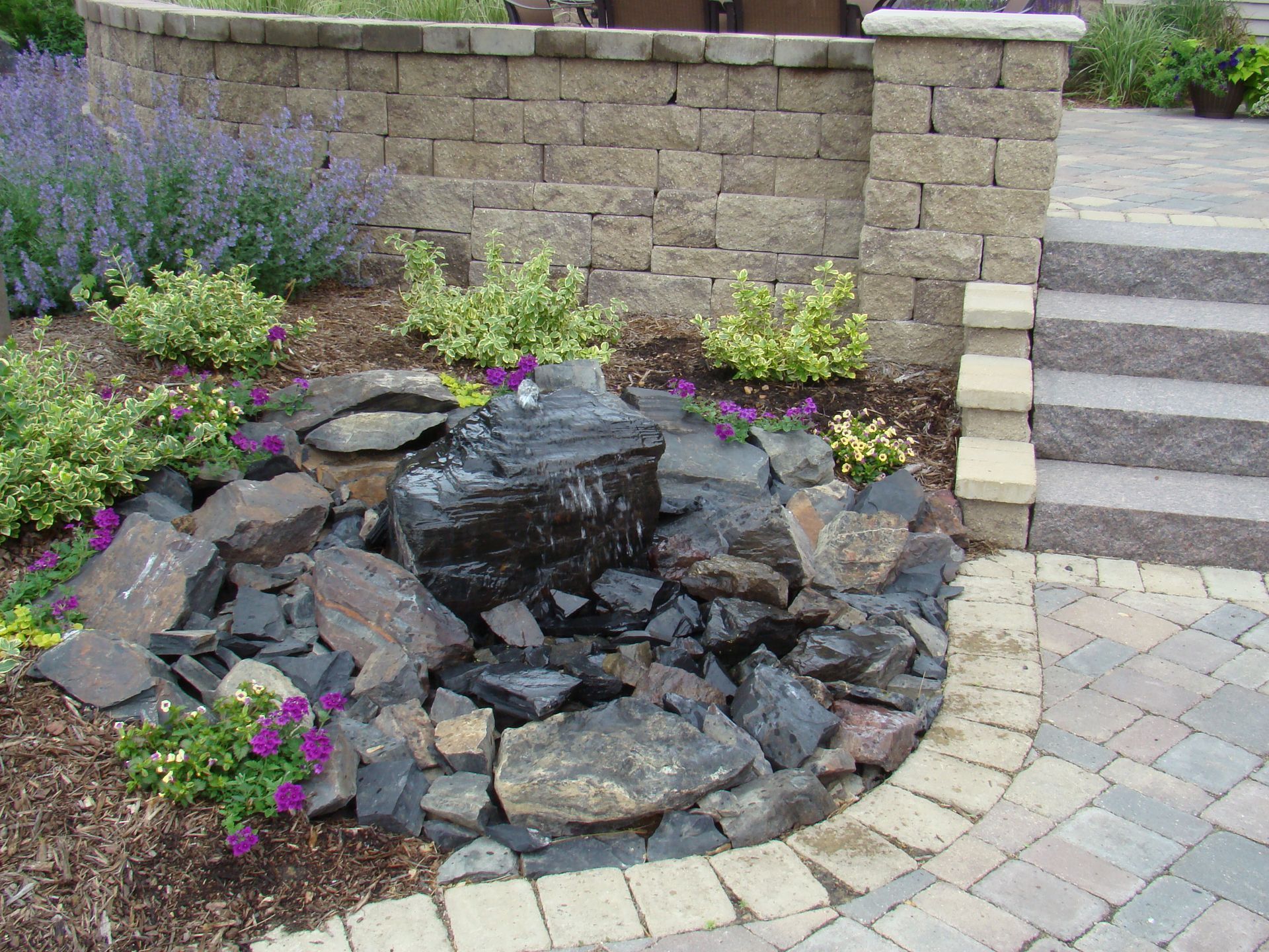 Water fountain feature in a garden, surrounded by dark rocks and purple flowers.