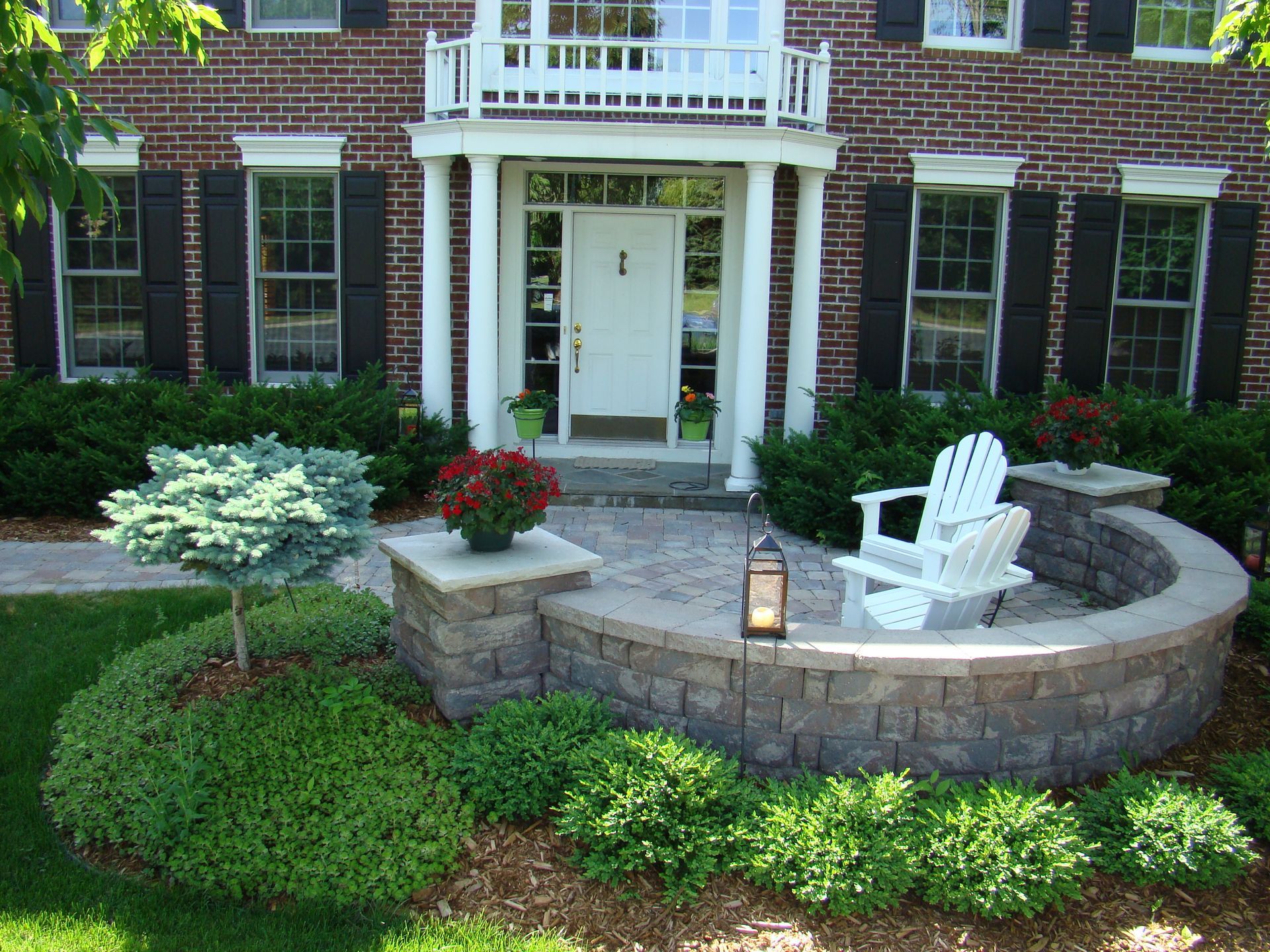 Brick house with front porch and patio seating area, shrubs, and a lantern.