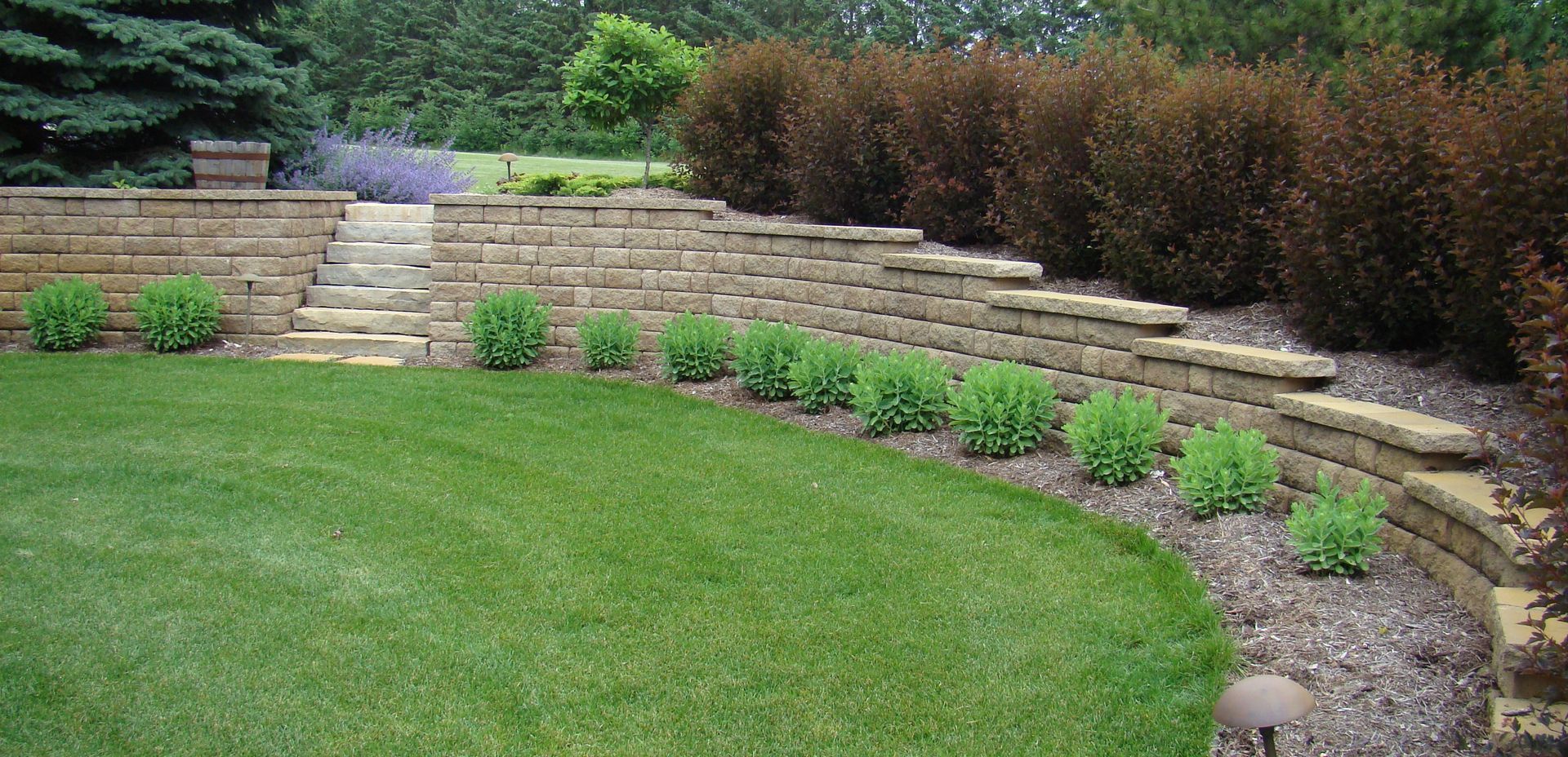 A tiered landscape with stone retaining walls, green lawn, steps, and shrubs.