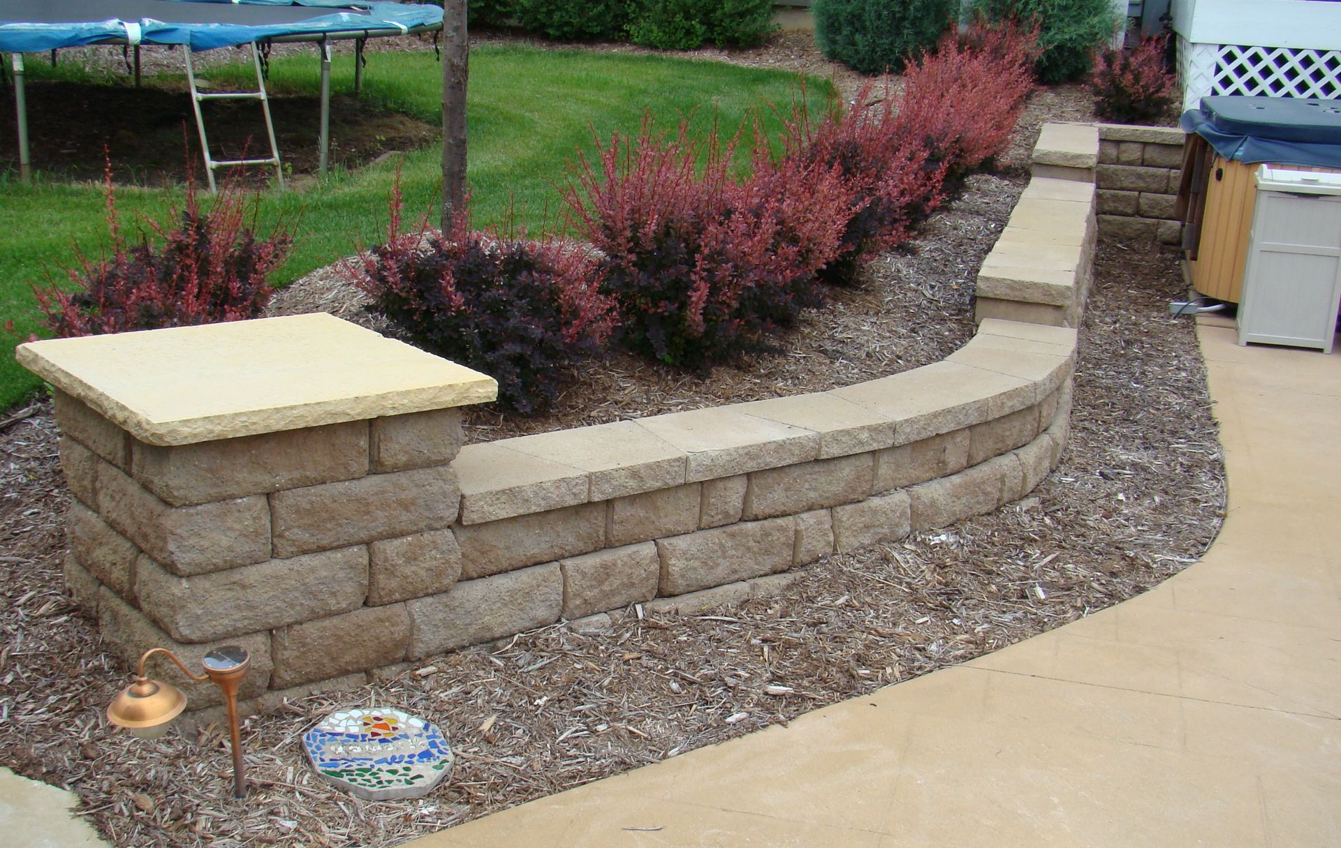 Stone retaining wall with red bushes, brown mulch, and beige concrete.