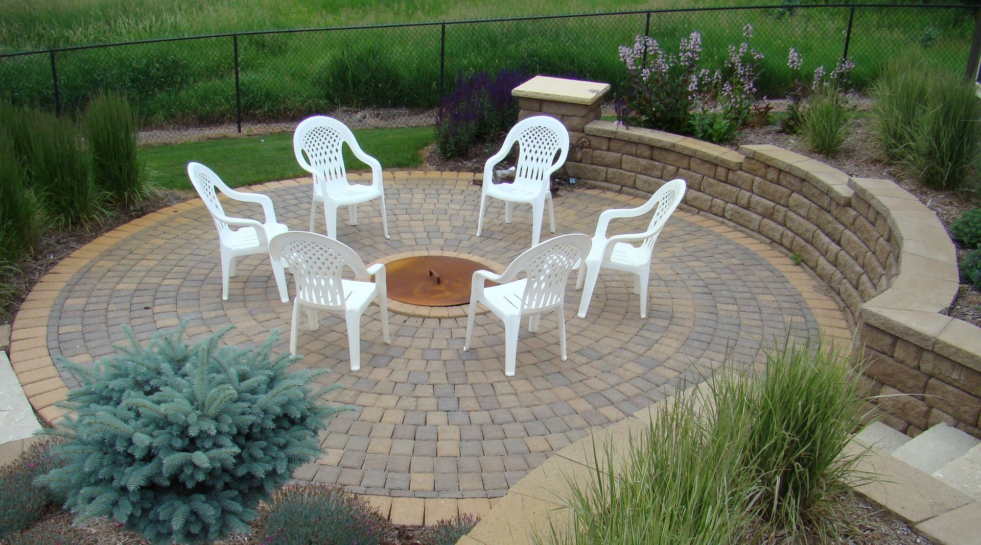 Circular brick patio with fire pit, white chairs, and tiered retaining wall.