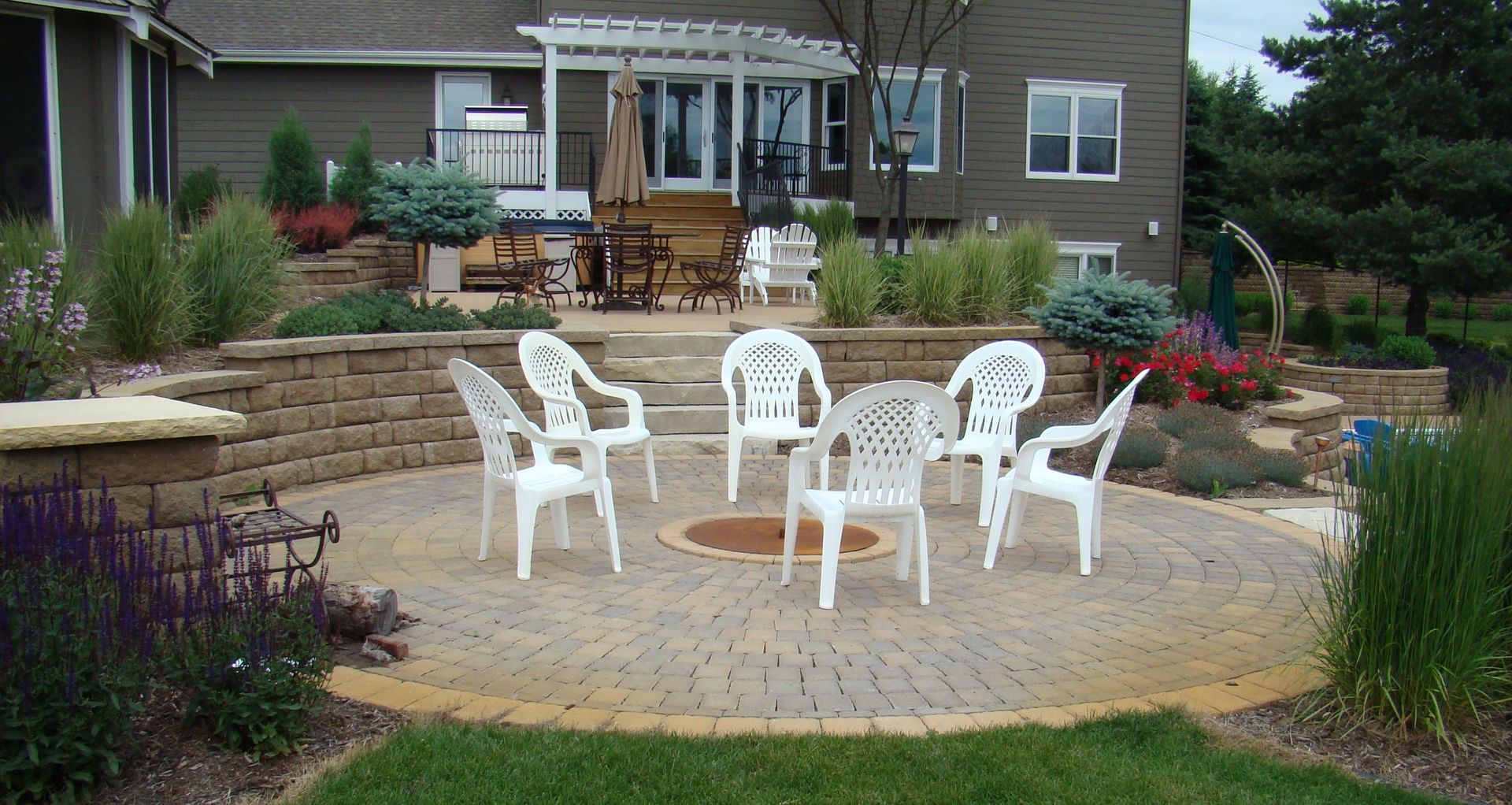 Backyard patio with fire pit surrounded by white chairs; house in background.