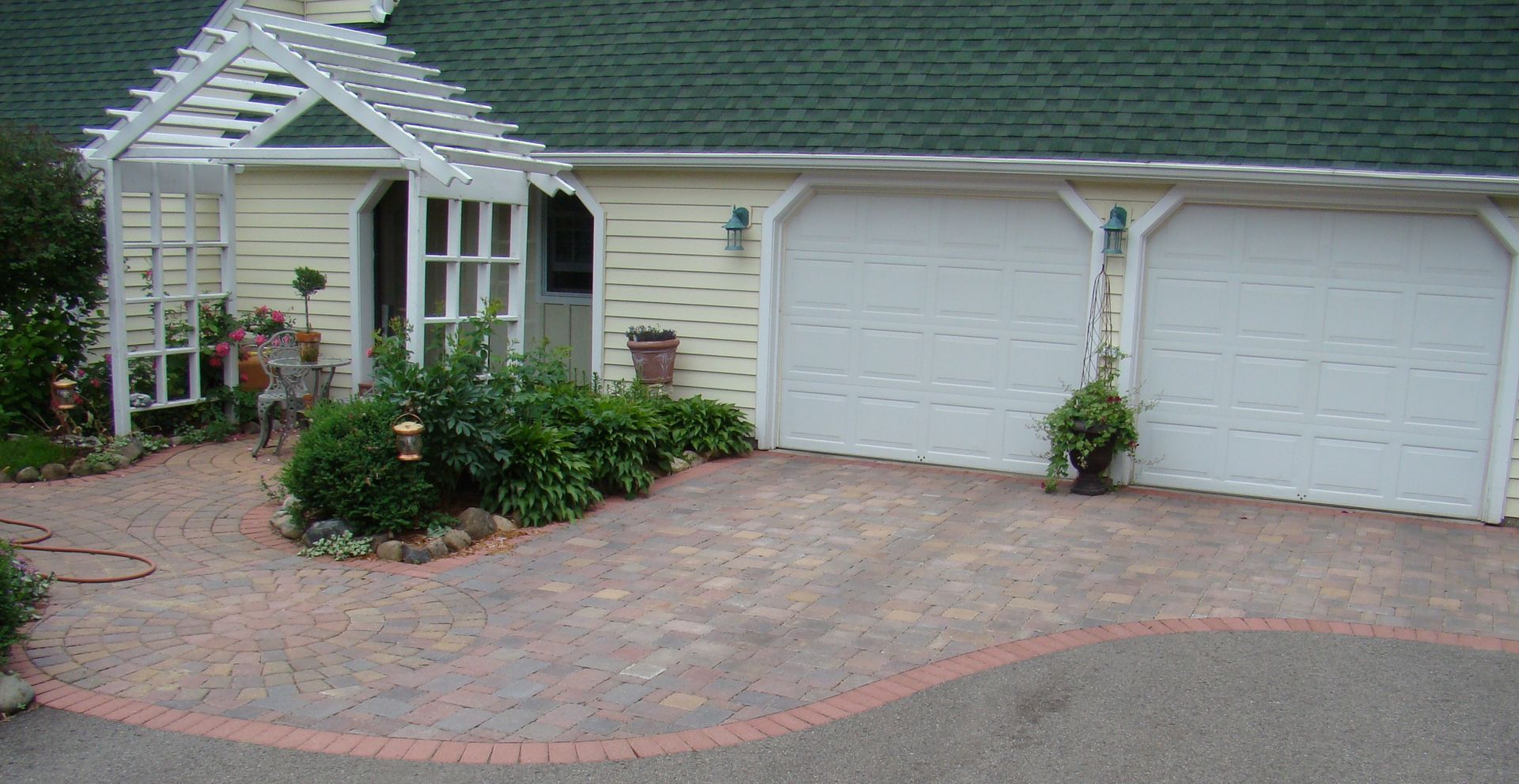 Garage with white doors and a brick driveway, light yellow exterior, and a green roof.