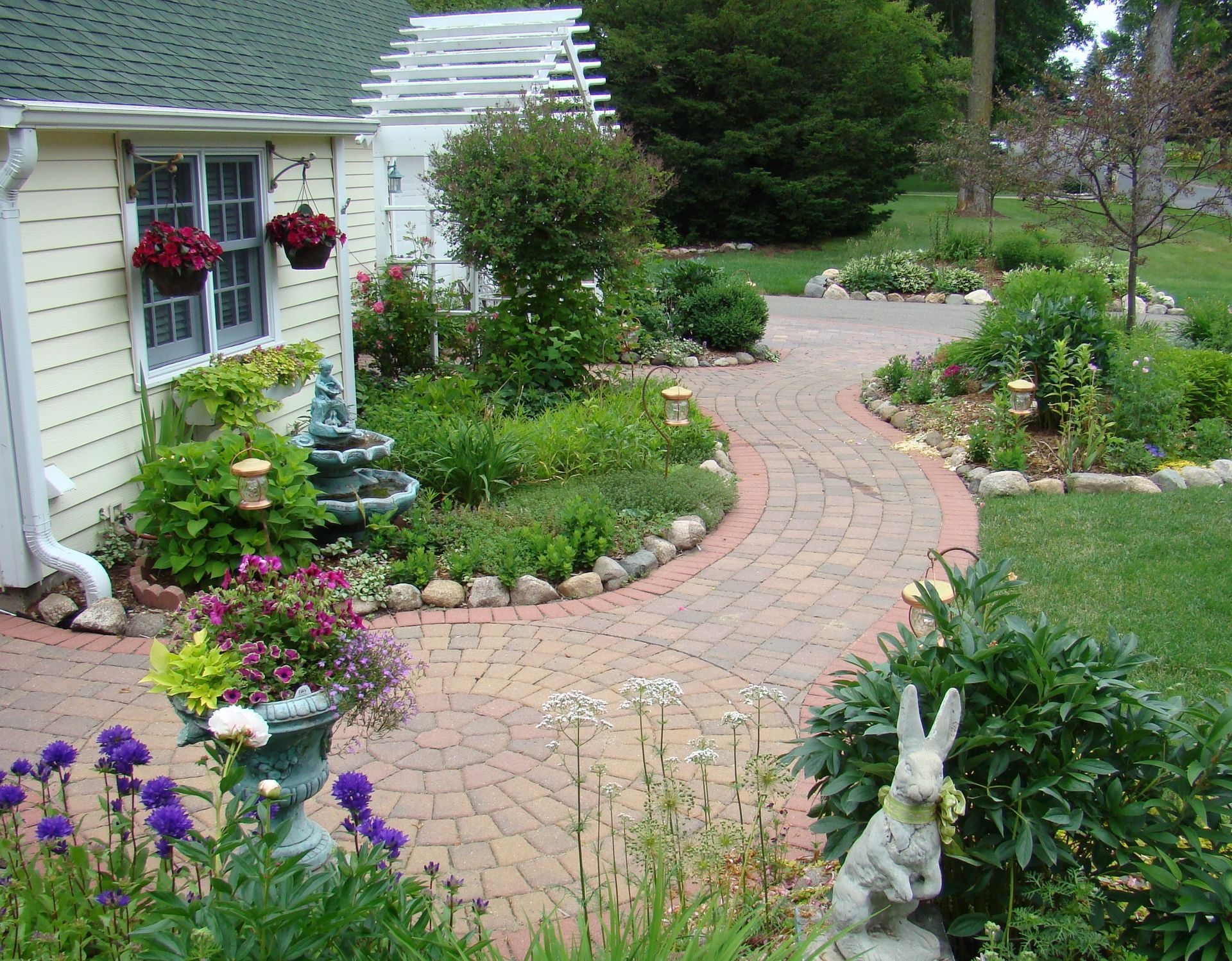 Brick pathway winds through a colorful garden with flowers, a fountain, and a small building.