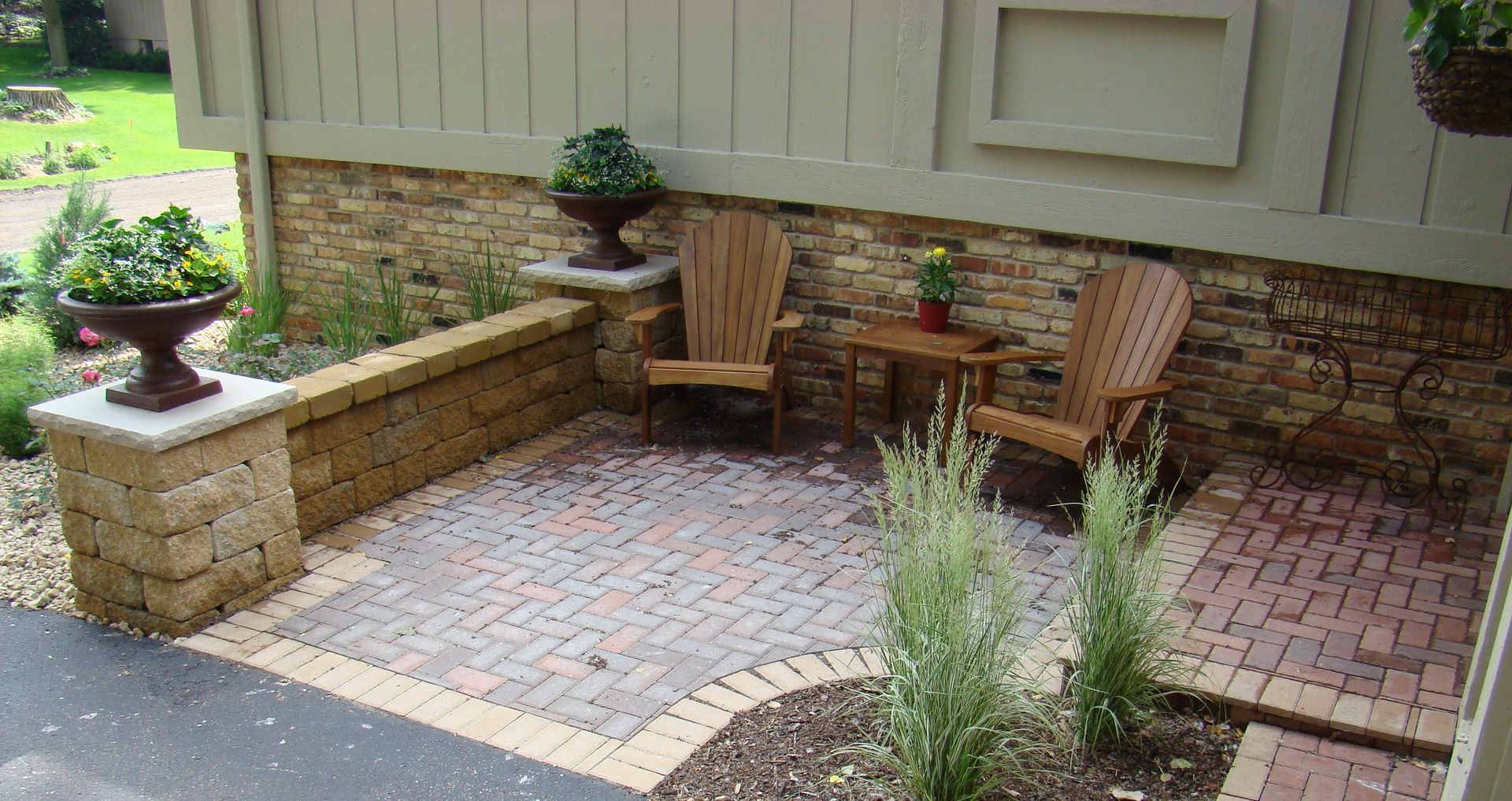 Patio with brick pavers, two wooden chairs, small table, stone wall, and potted plants.