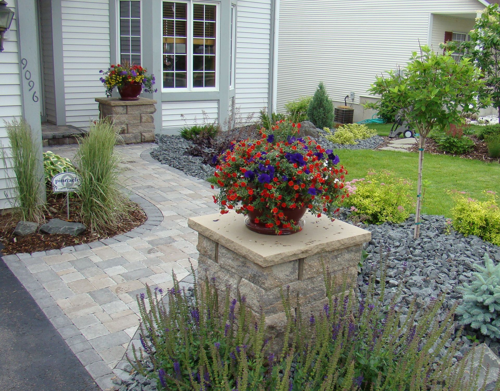 Stone walkway leading to a house entrance with flower pots on pillars, lush greenery, and a garden.