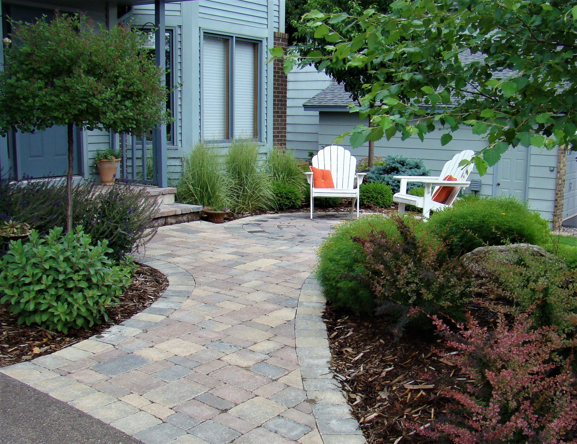 Brick pathway leads to white Adirondack chairs in a garden setting, with shrubs and trees.