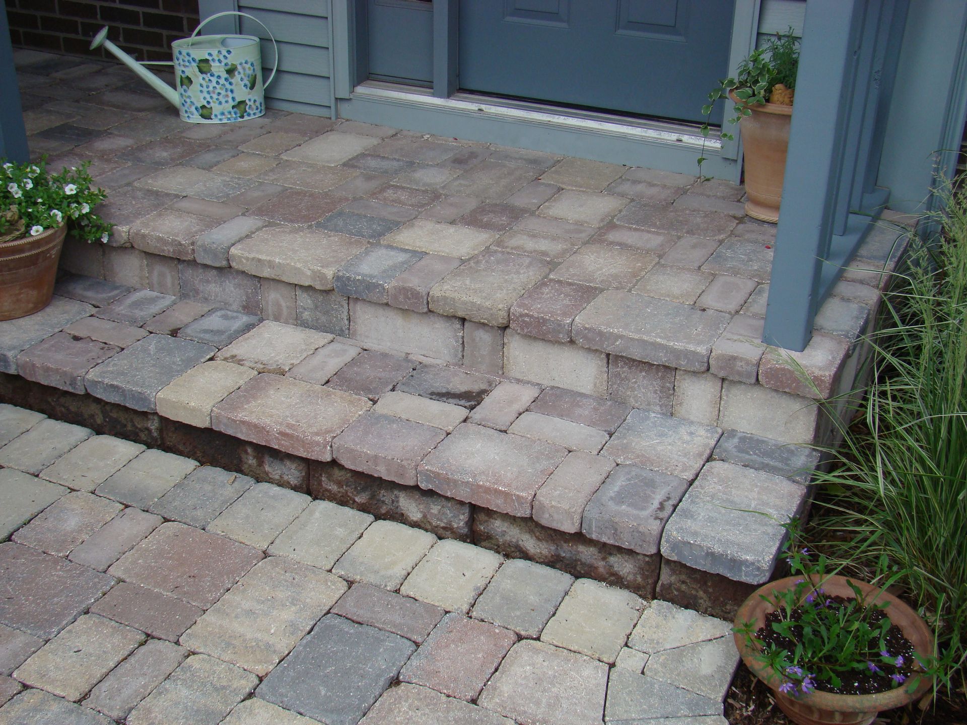 Stone brick steps leading to a gray door, with potted plants on the landing and a watering can nearby.
