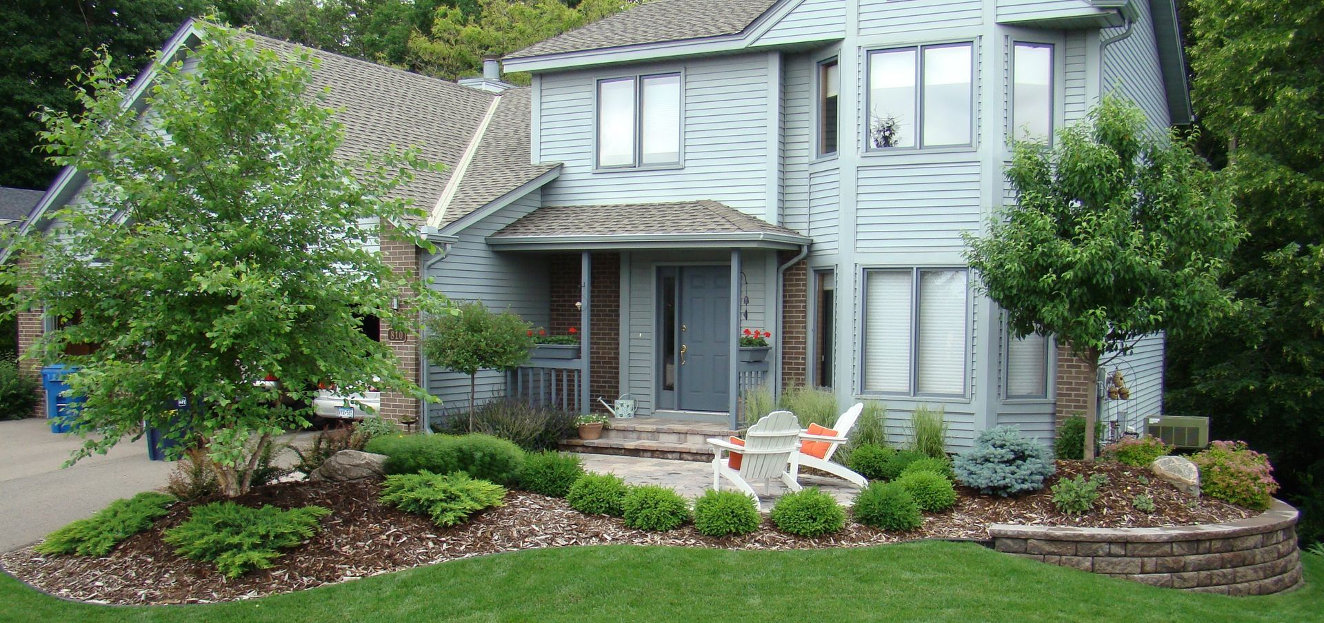 A two-story blue house with landscaping, a stone retaining wall, and green lawn.