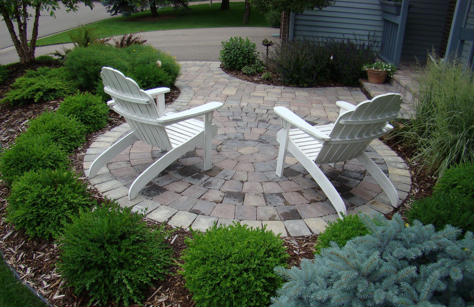 Two white Adirondack chairs on a brick patio surrounded by green shrubs, in a garden.