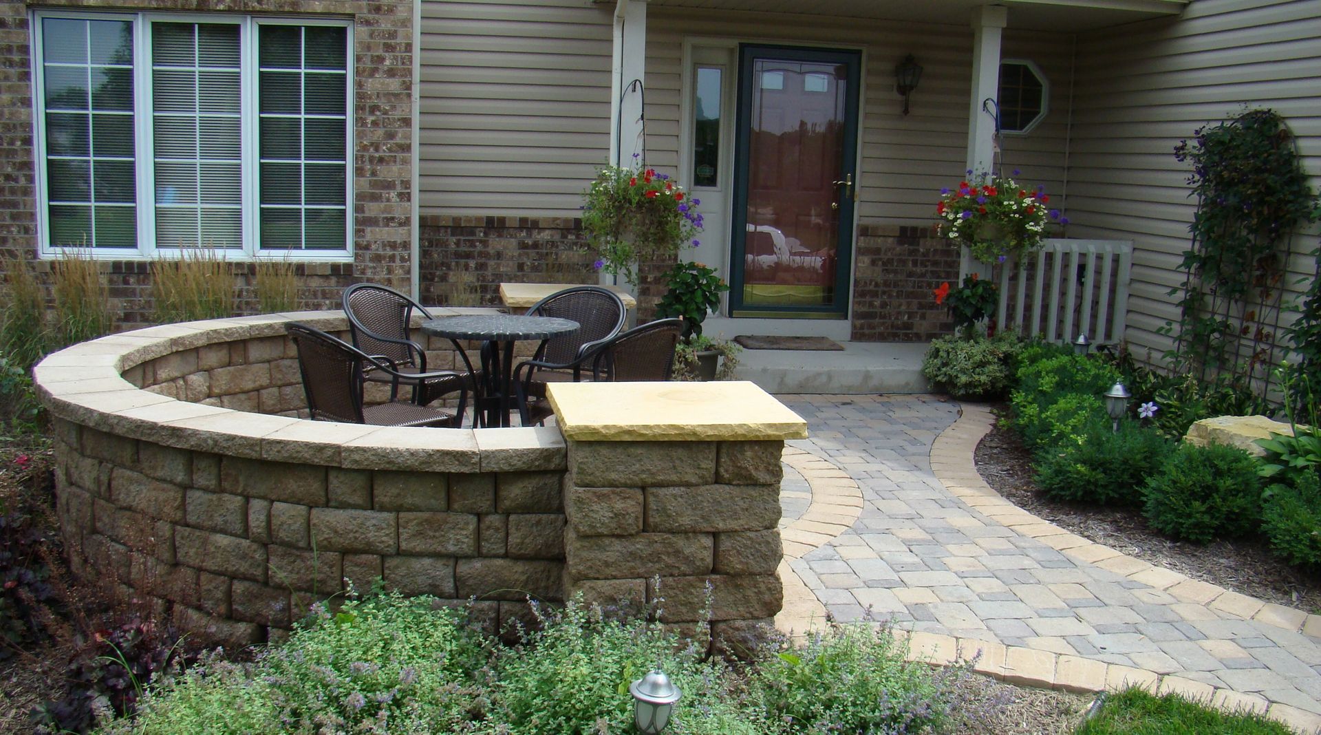 Outdoor patio with a curved stone wall and table with chairs. A brick pathway leads to a front door.
