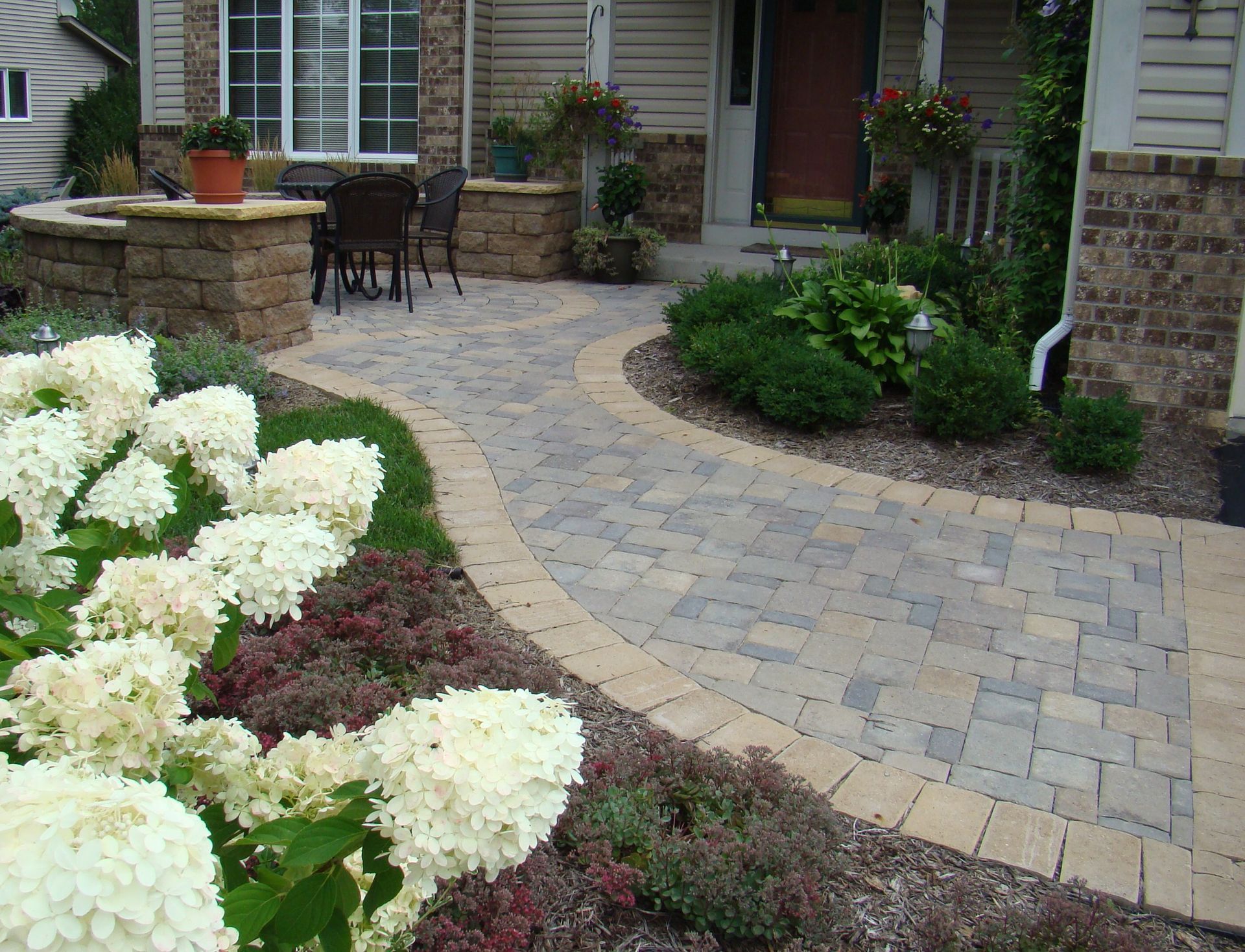 Stone pathway leads to a house with a curved design, bordered by plants and flowers.
