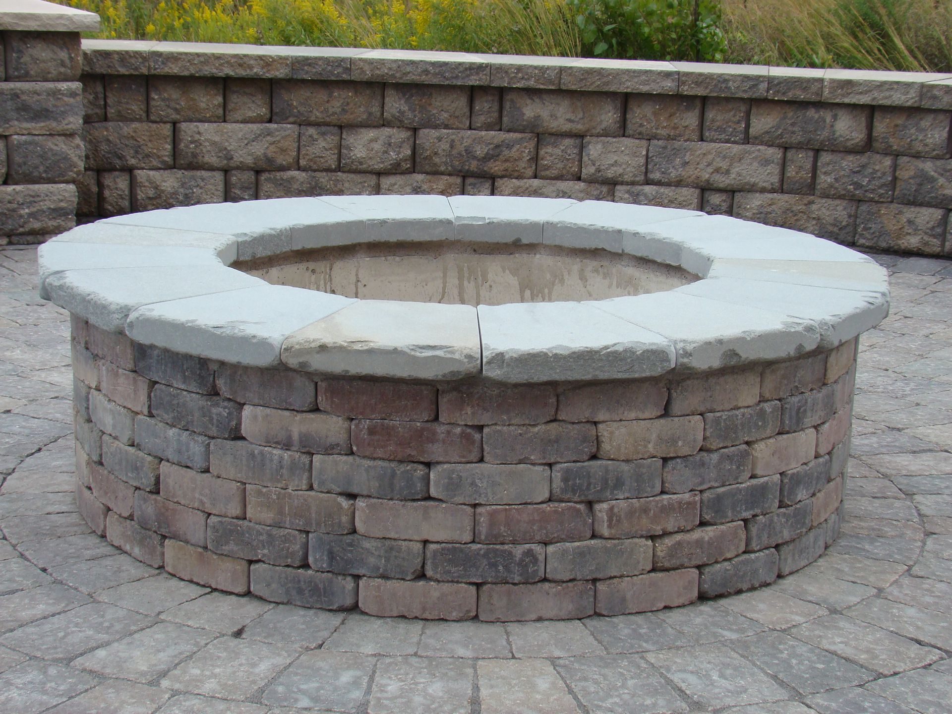 Circular brick fire pit with stone coping, on a paved patio, with a brick wall backdrop.