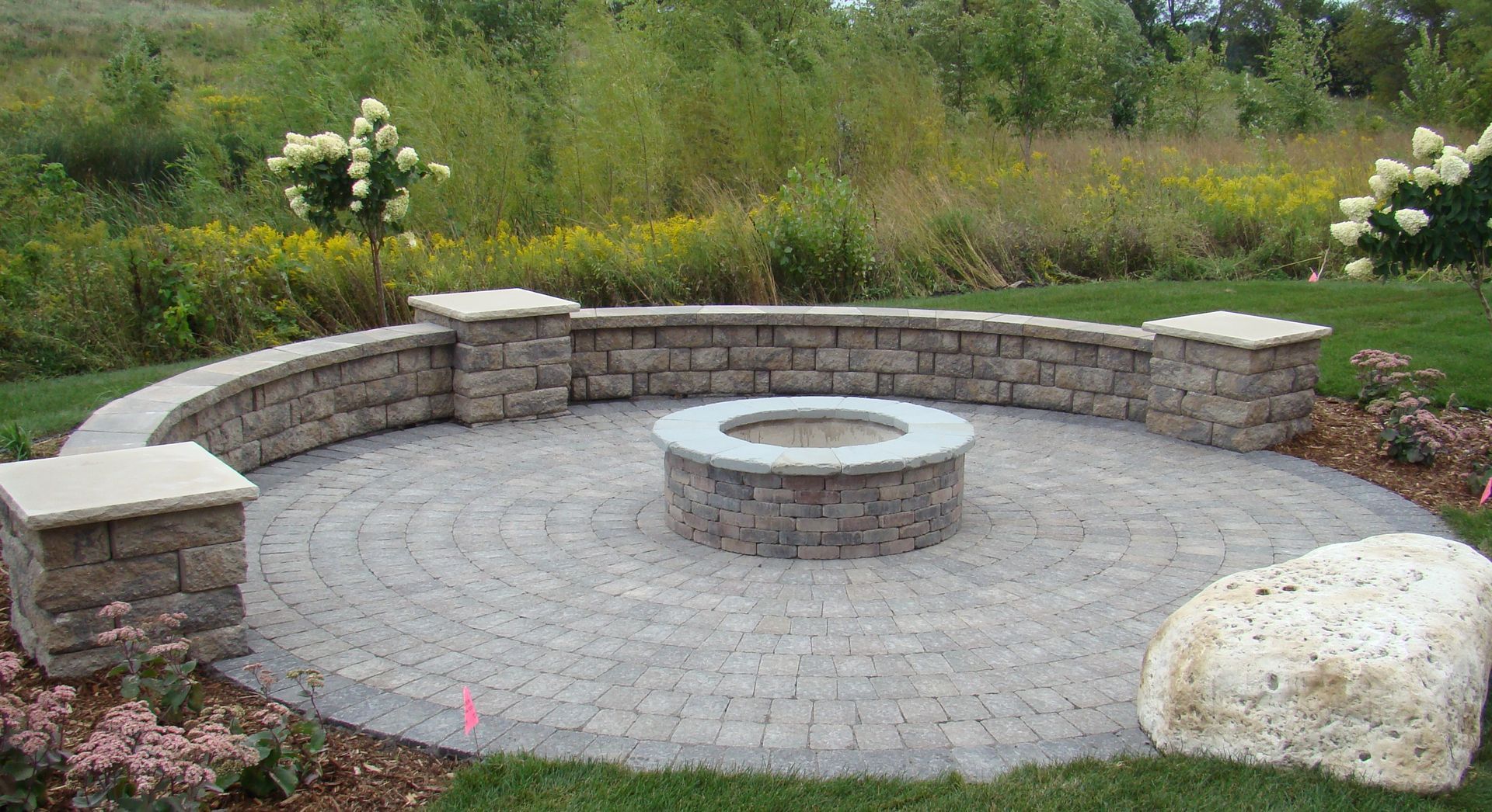 Circular stone patio with fire pit and curved wall in a grassy outdoor setting.