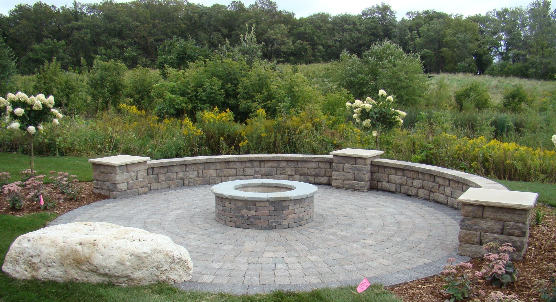 Circular stone patio with fire pit and seating wall, surrounded by garden and trees.