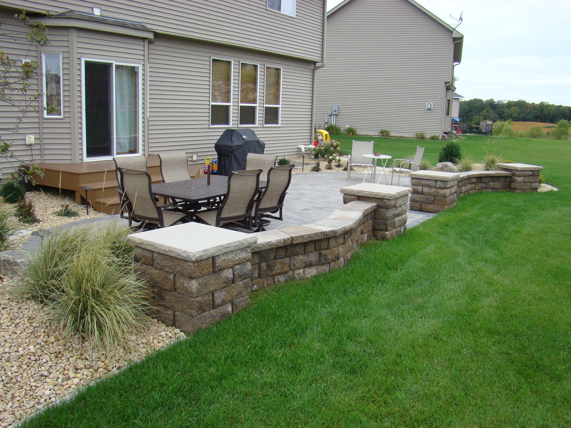 Backyard patio with dining set, grill, and low stone wall. Green lawn and beige siding.